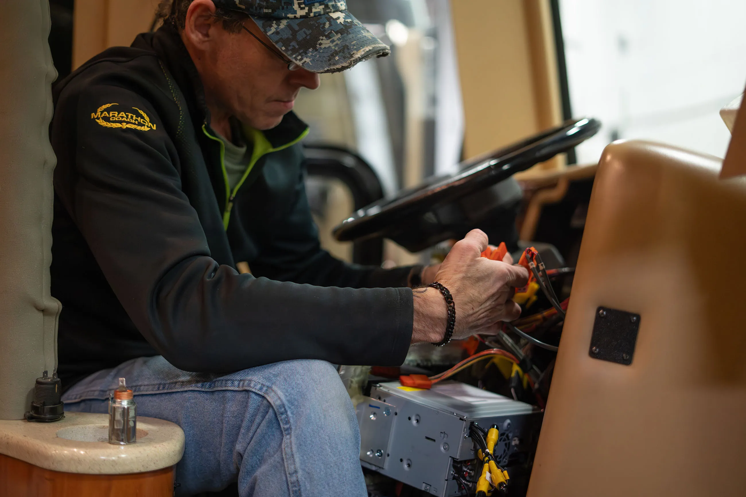 Man in a camo cap and glasses fixing car stereo wiring inside a vehicle.