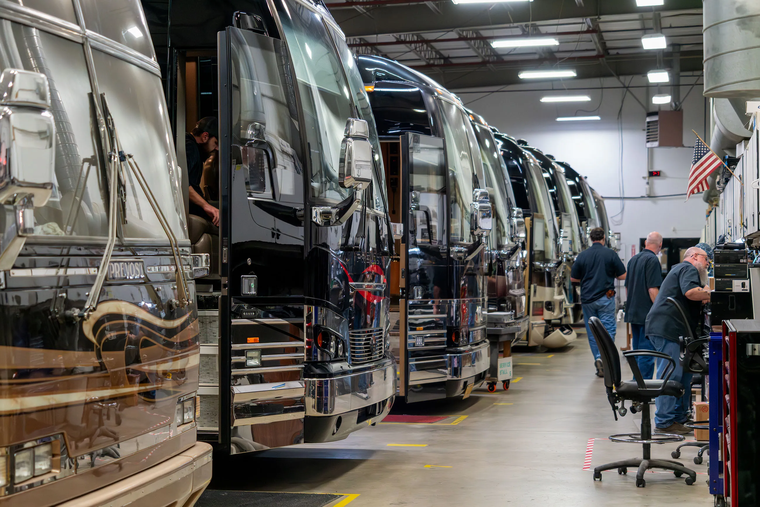 Row of large luxury buses in a well-lit service garage with technicians working and an American flag displayed.