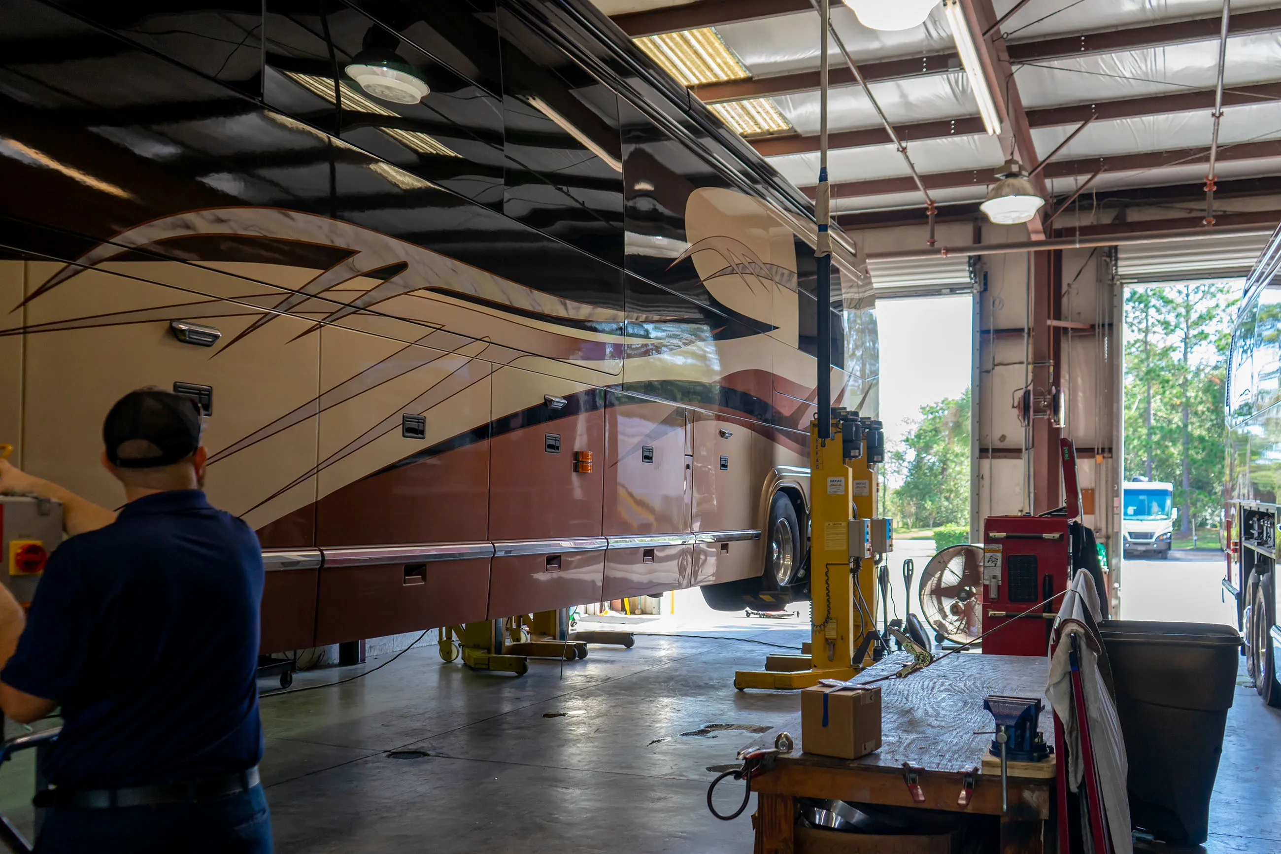 Mechanic operating controls with motorhome lifted on hydraulic vehicle lift inside a service garage.