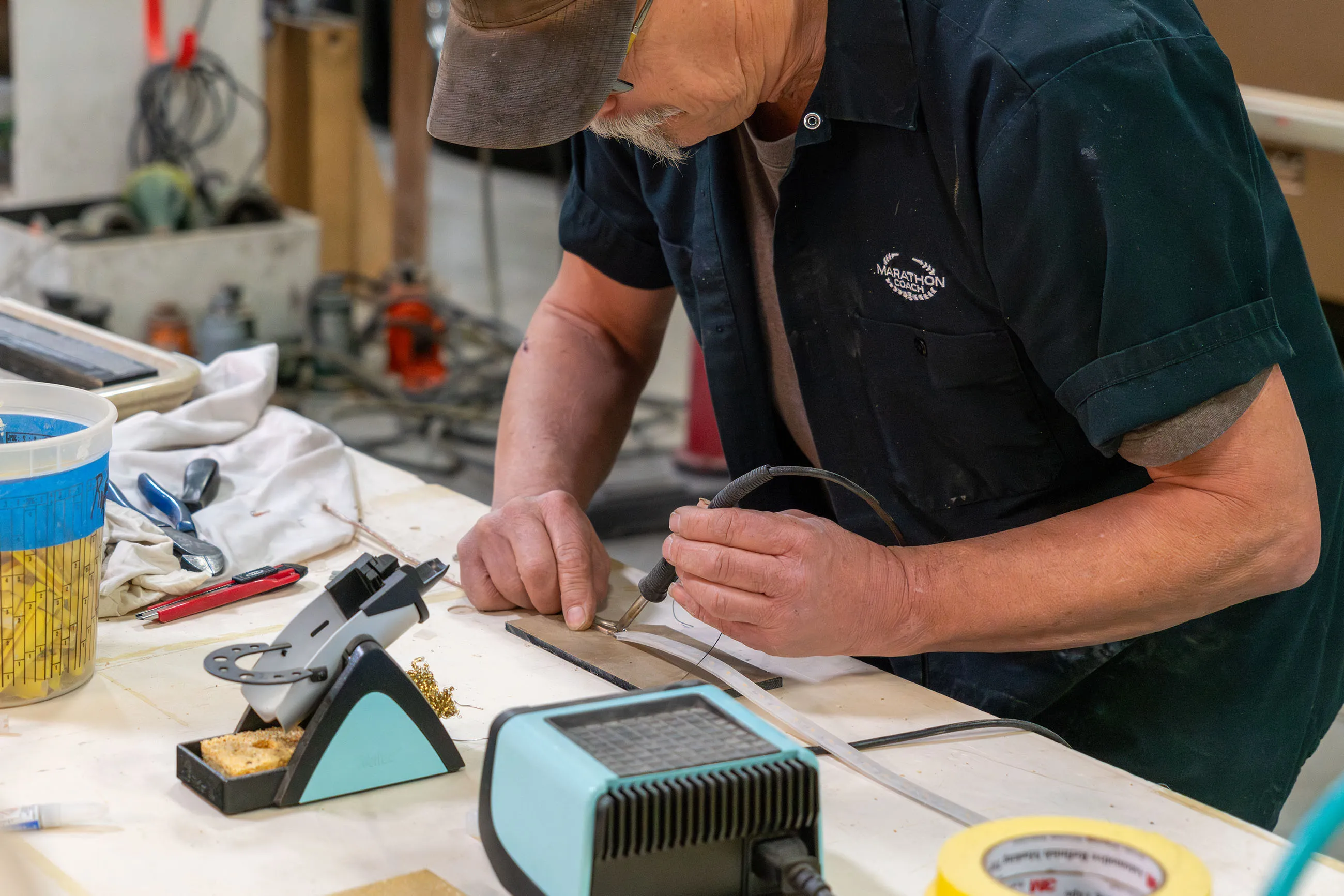 Man in a dark shirt using a soldering iron to work on a small metal component at a cluttered workbench.