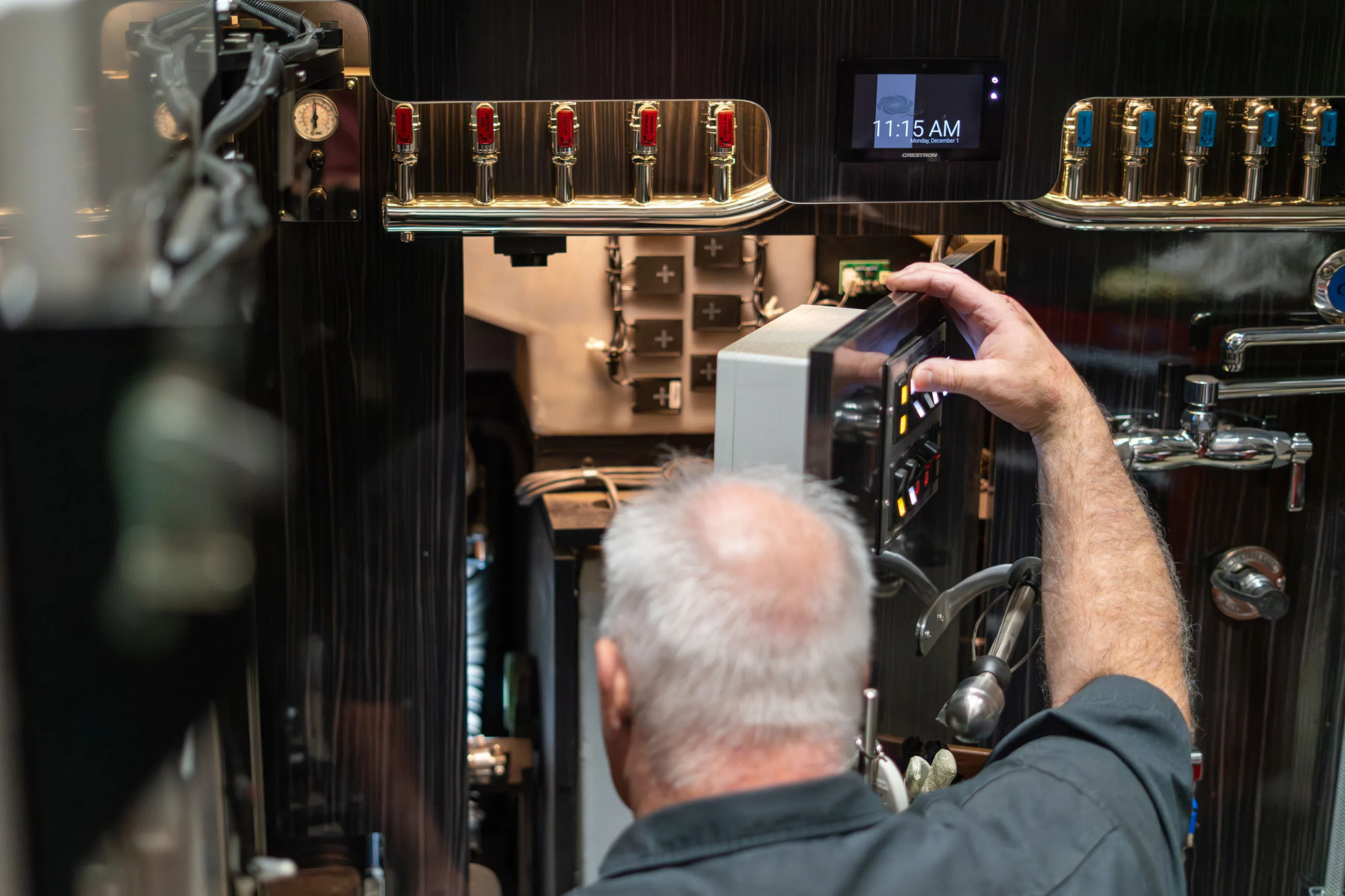 Man adjusting control panel inside a polished metal brewing or beverage dispensing machine with taps and a digital display showing 11:15 AM.