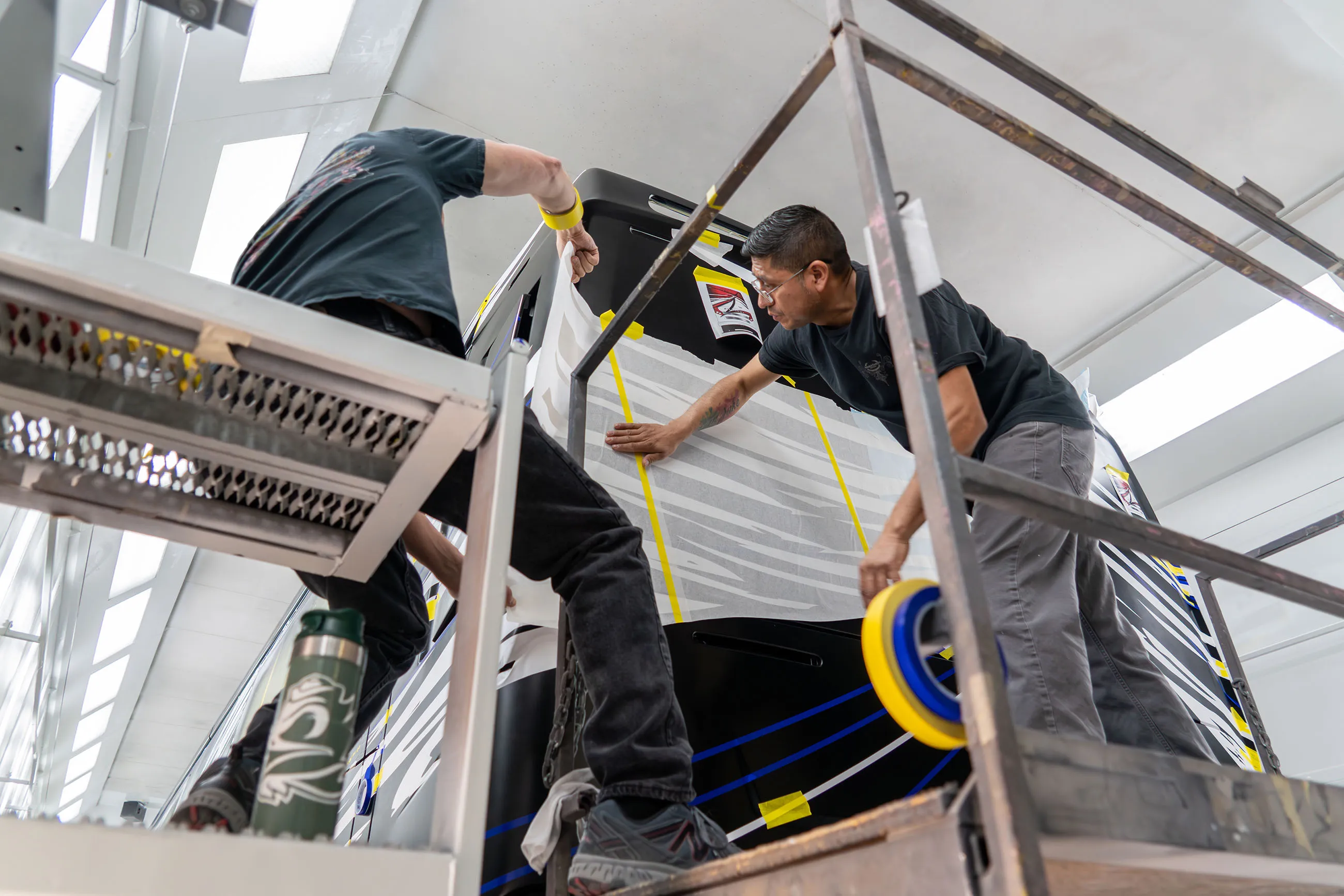 Two men applying a large adhesive graphic to a black surface, working on scaffolding indoors.