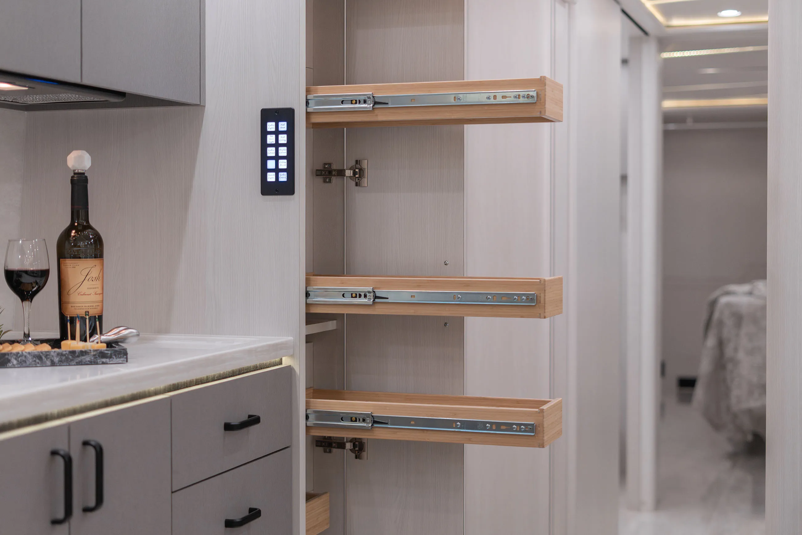 Modern kitchen interior showing open pull-out drawers in a light wood cabinet and a bottle of red wine with a glass on the countertop.