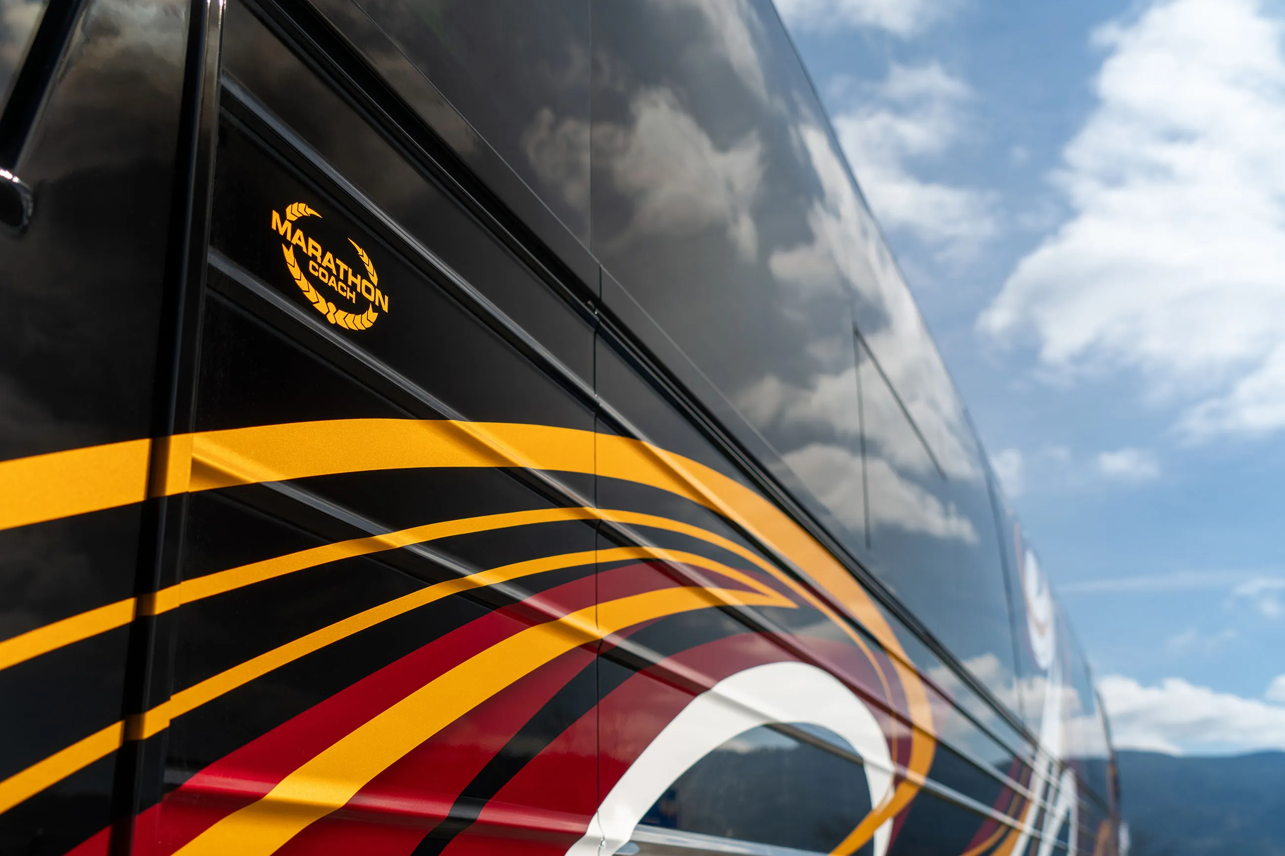 Side view of a black Marathon Coach vehicle with yellow, red, and white swoosh designs under a partly cloudy sky.