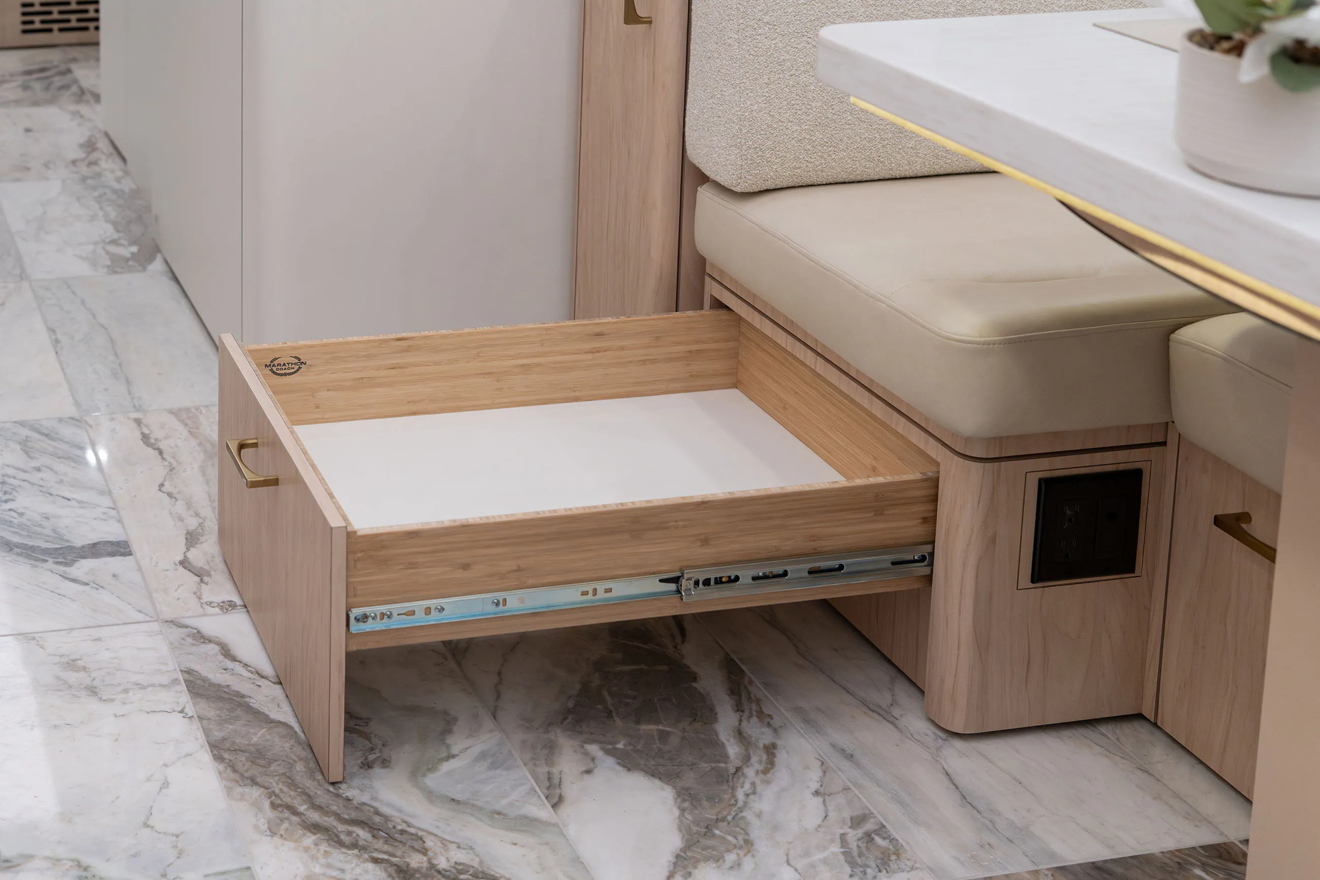 Open wooden drawer with metal slides underneath beige cushioned bench seating on marble floor.