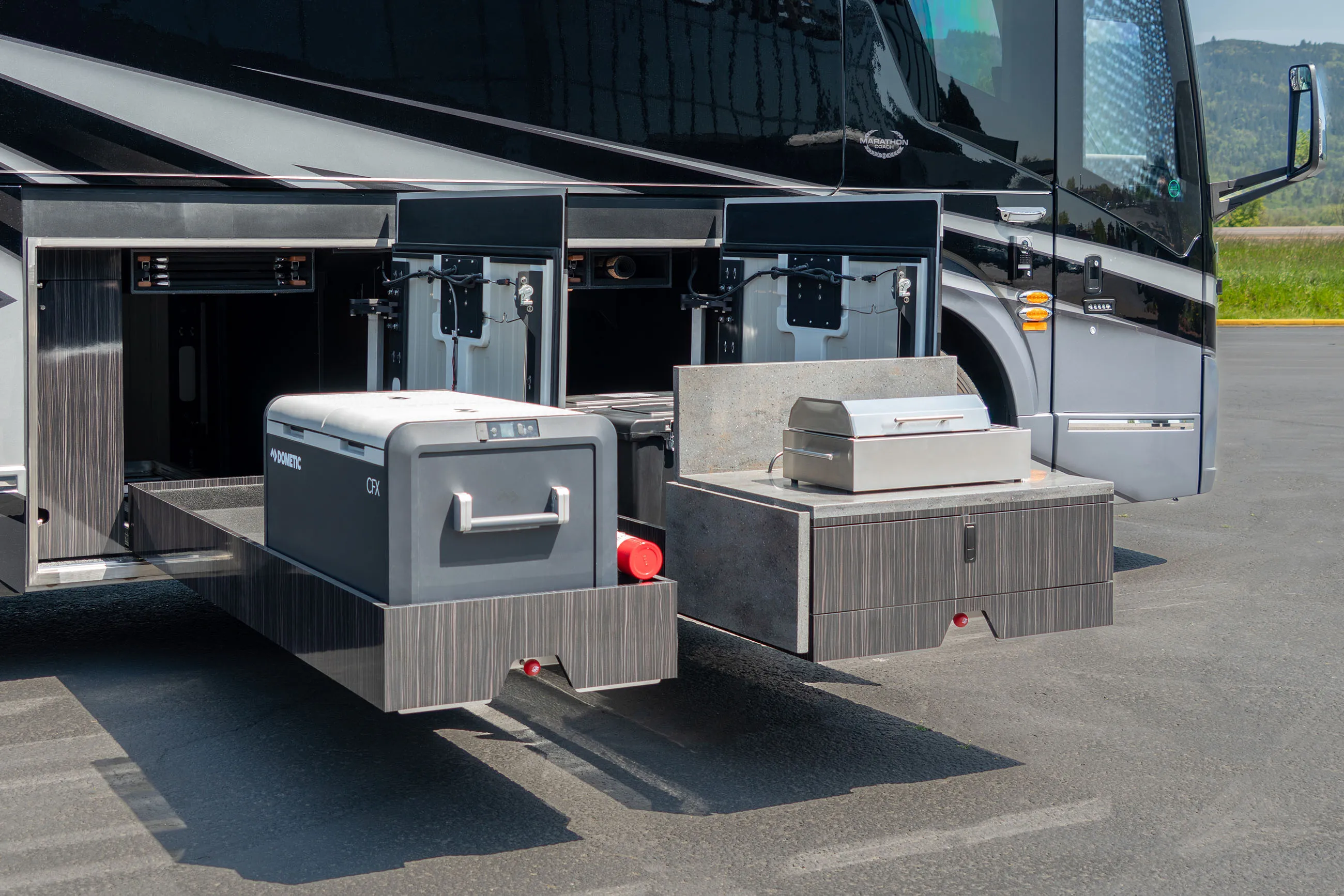 Open side compartments of a motorhome revealing two slide-out trays, one with a portable fridge and the other with a closed stainless steel grill.