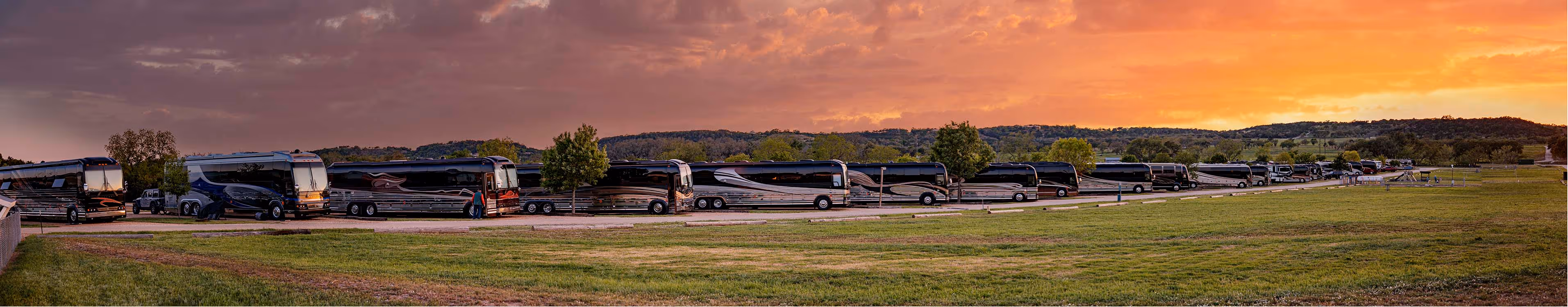 Long row of black luxury tour buses parked on a paved lot at sunset with pink and orange sky and distant trees and hills.