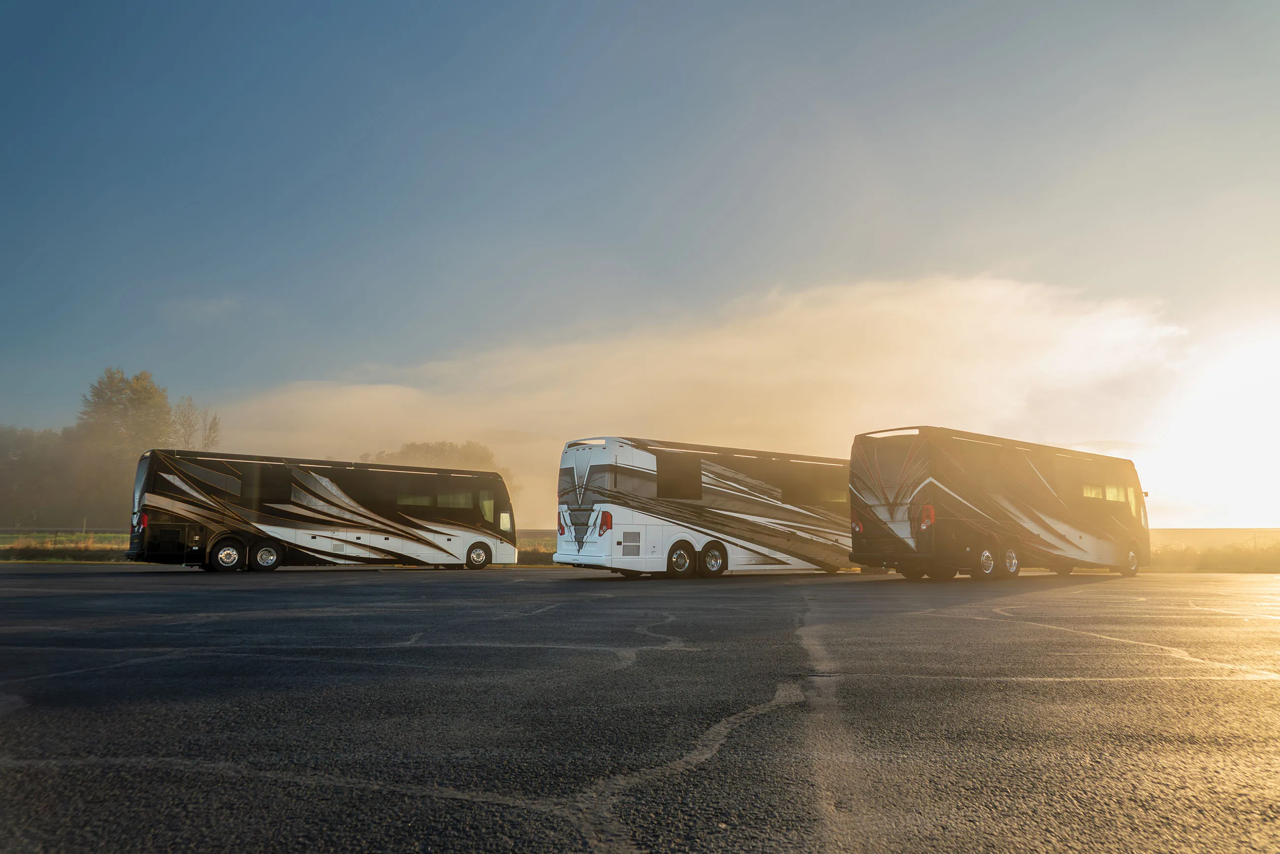Three luxury motorhomes with black, white, and silver designs parked on an empty lot at sunrise with a clear sky.