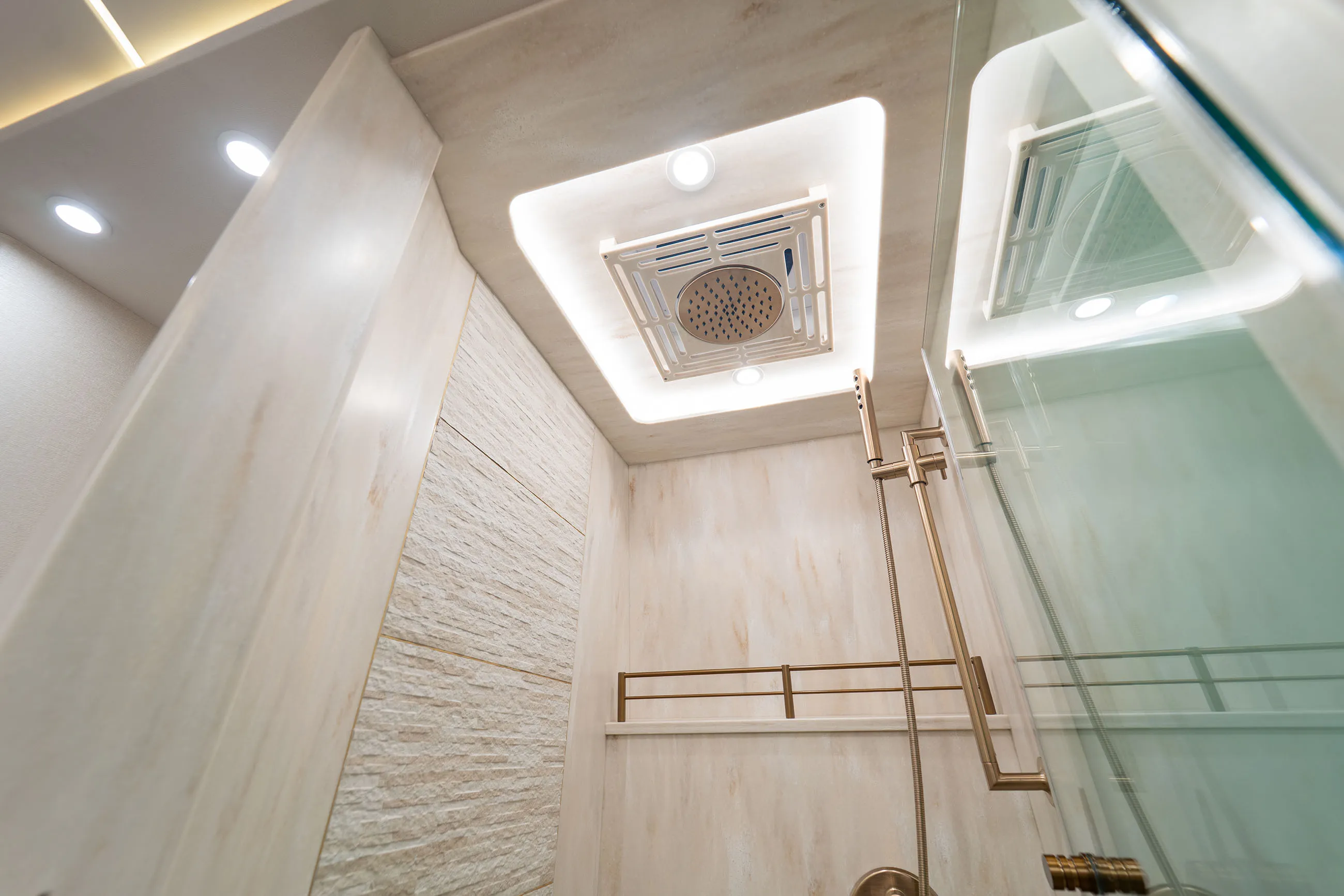 Modern shower interior with beige stone walls, ceiling-mounted rainfall showerhead, handheld shower, and glass door.