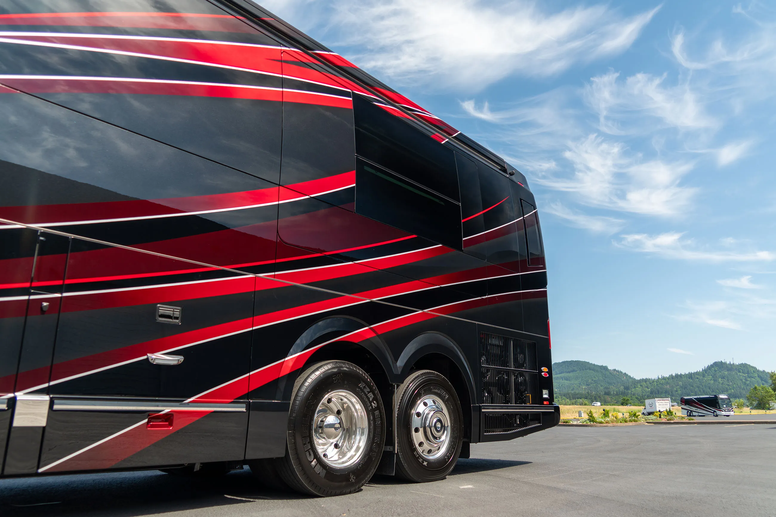 Black and red luxury coach bus parked on a road with trees and hills in the background under a partly cloudy sky.