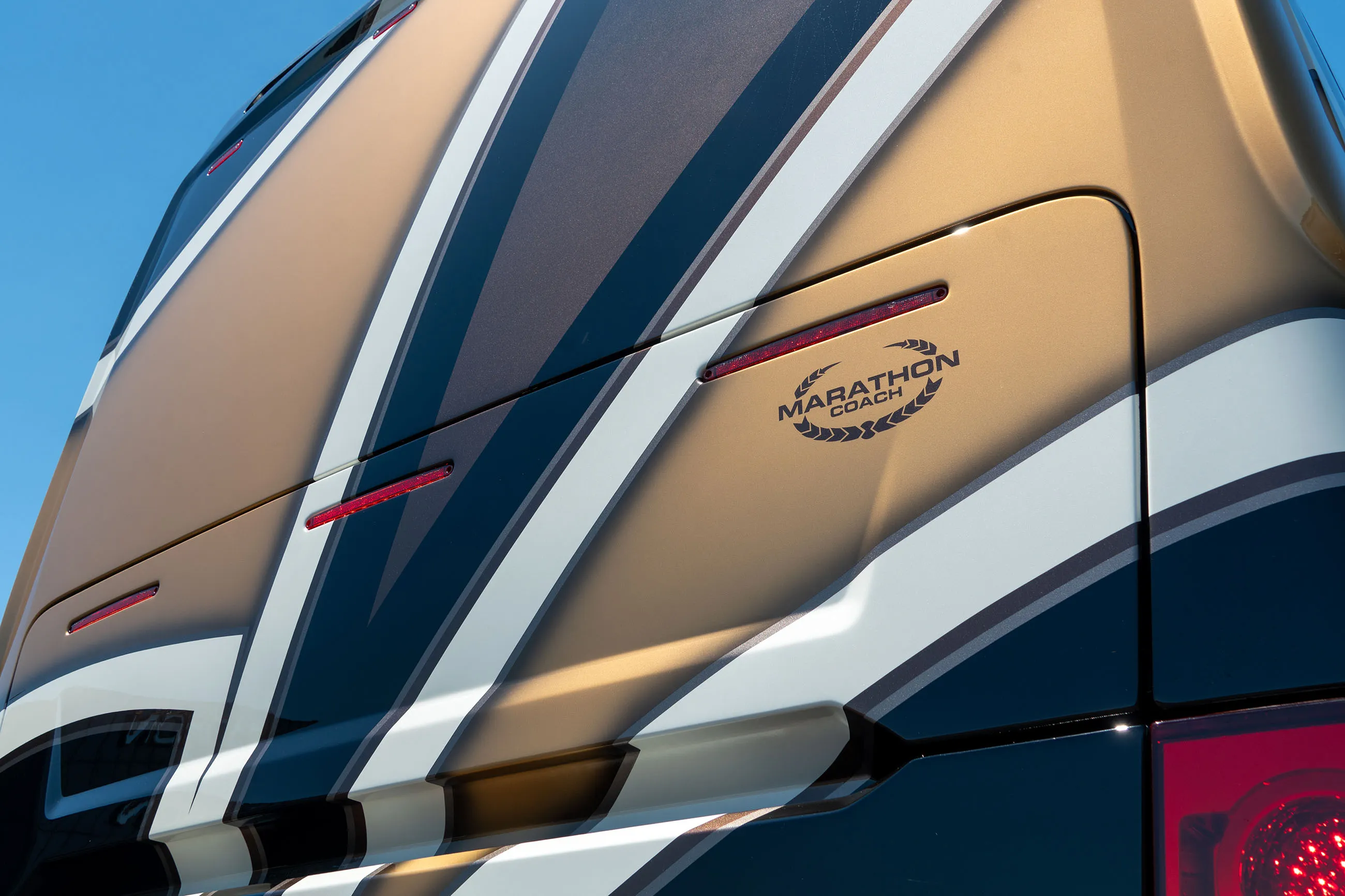 Close-up of the rear side panel of a Marathon Coach RV with gold, black, and white stripes and red lights.