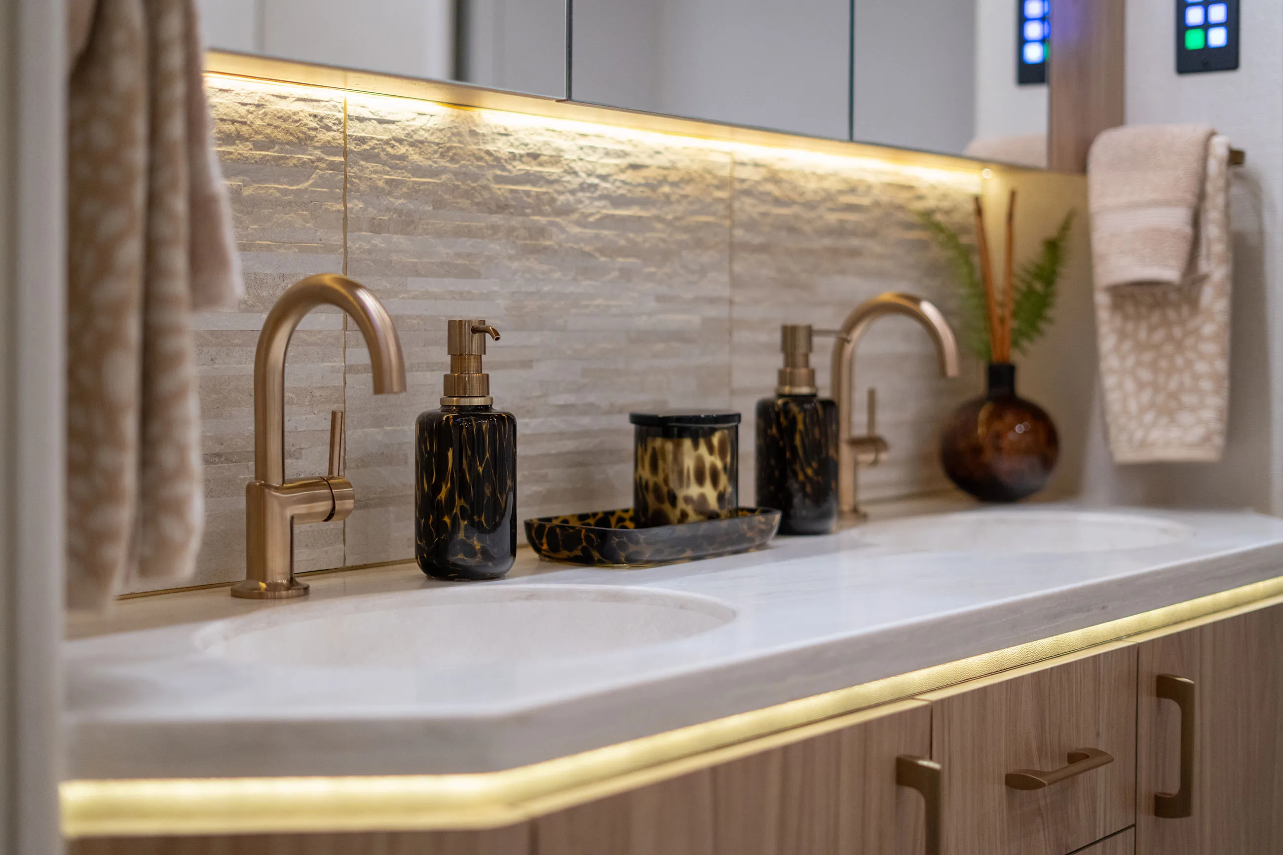 Modern bathroom double sink vanity with gold faucets, patterned soap dispensers, candle, and beige towels.