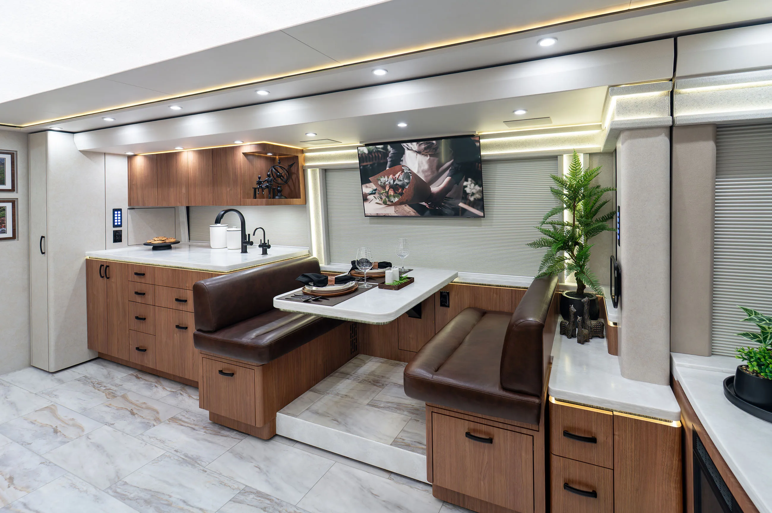 Modern RV interior with a kitchenette, leather bench seating, a dining table set for two, and a wall-mounted TV.