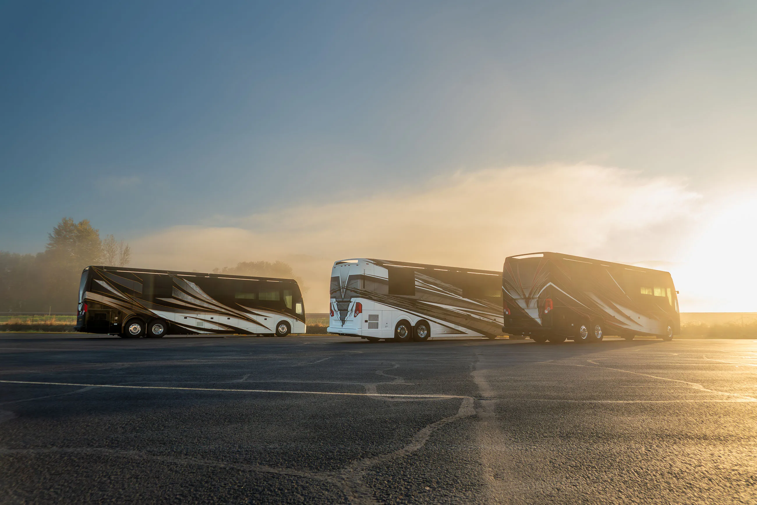 Three luxury motorhomes parked side by side on an asphalt surface during a foggy morning with sunlight breaking through.