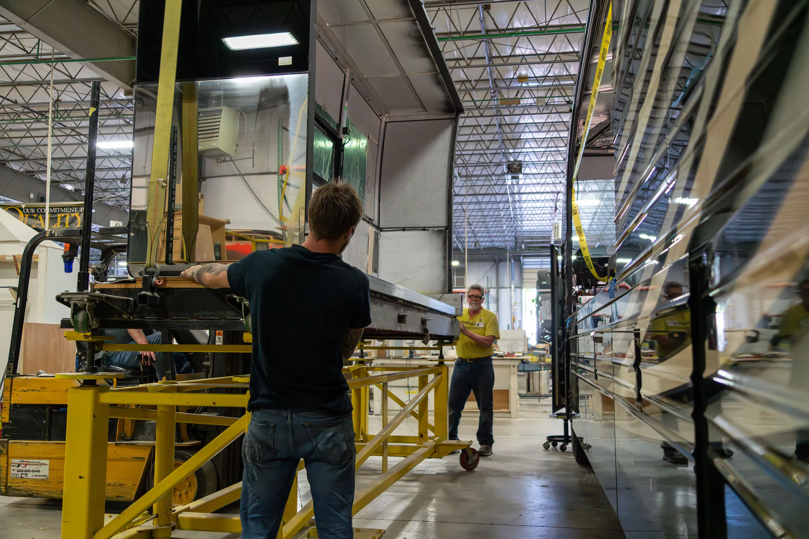 Two men handling a large reflective metal panel inside a spacious industrial workshop with machinery and equipment.