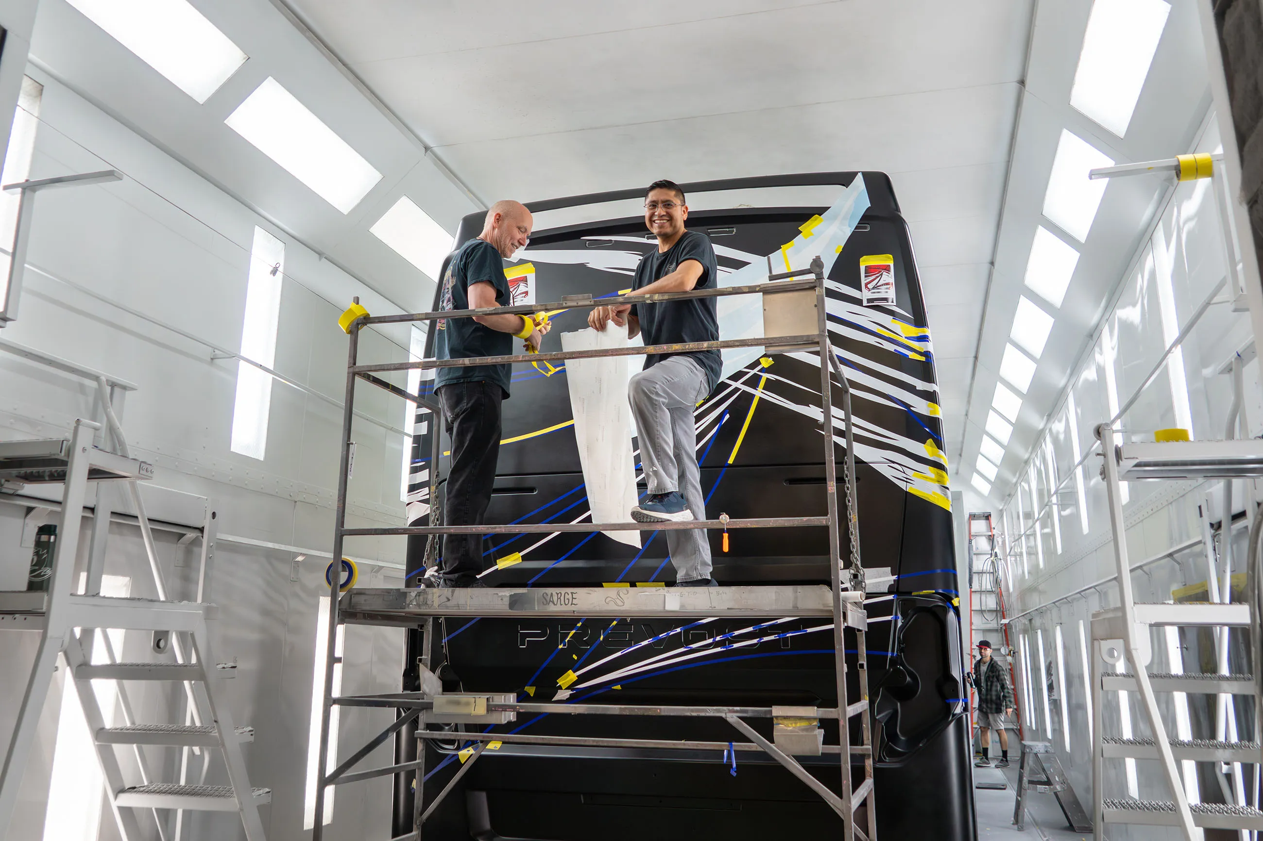 Two men on scaffolding applying white stencil vinyl to the rear of a large black vehicle with colorful geometric stripes in a well-lit workshop.