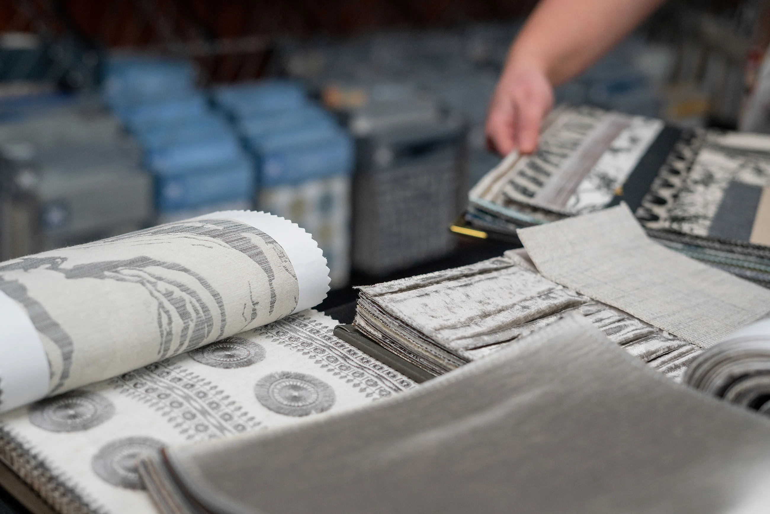 Close-up of various fabric upholstery samples with neutral tones and patterns spread on a table, a hand holding some swatches in the background.