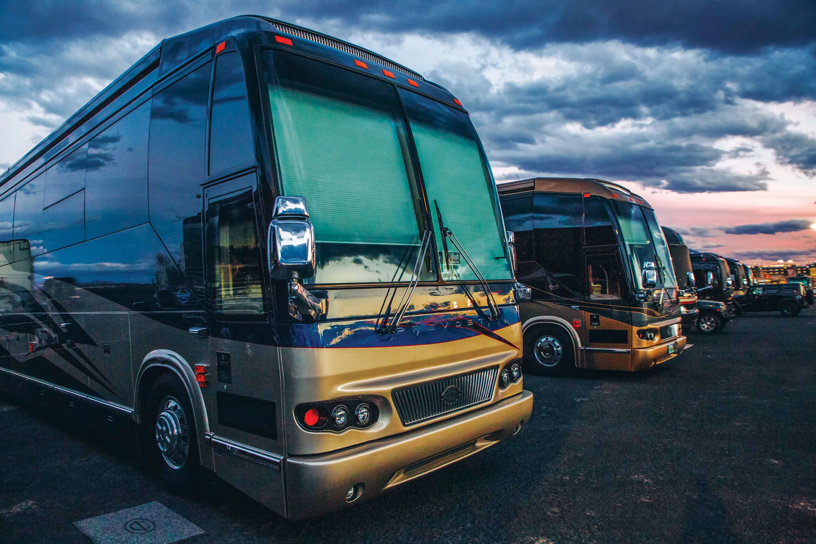 Row of luxury tour buses parked at dusk under a cloudy sky.