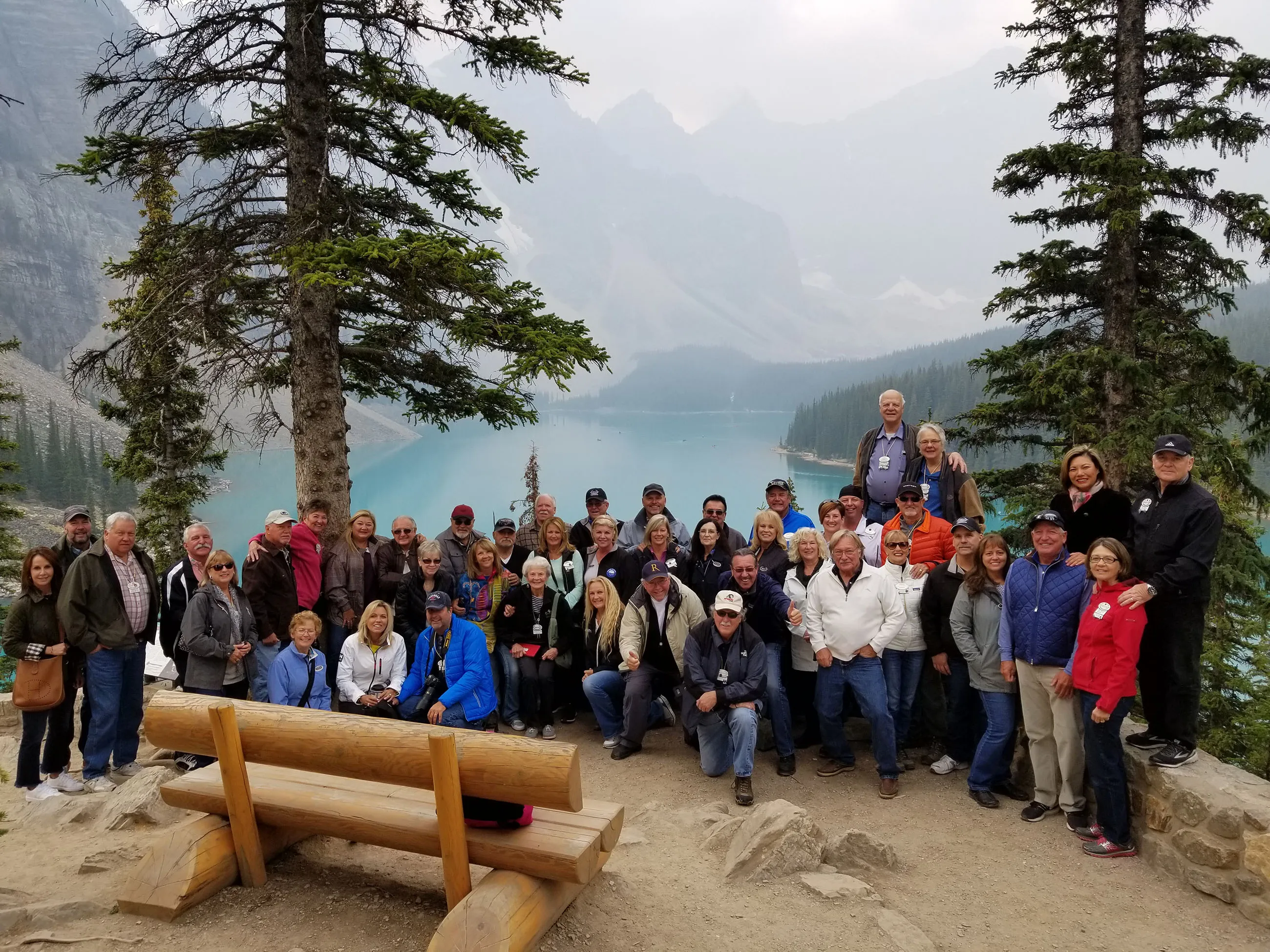 Large group of people posing together by a turquoise lake with mountains and trees in the background.