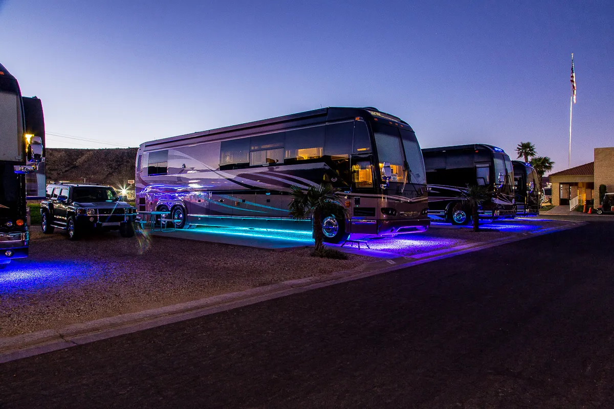 Luxury RVs parked at an RV park during dusk with blue and purple ambient lights underneath.