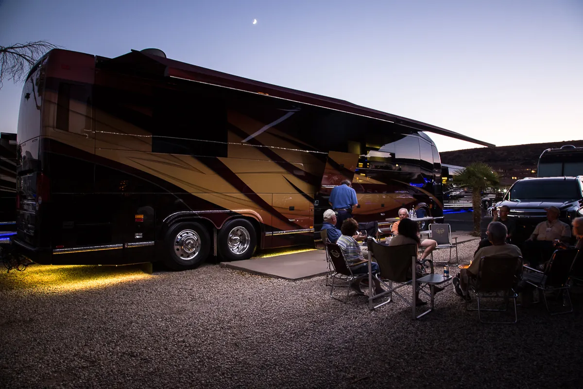 Group of people sitting on chairs around a table next to a large, brown and gold RV parked on gravel at dusk with a crescent moon in the sky.