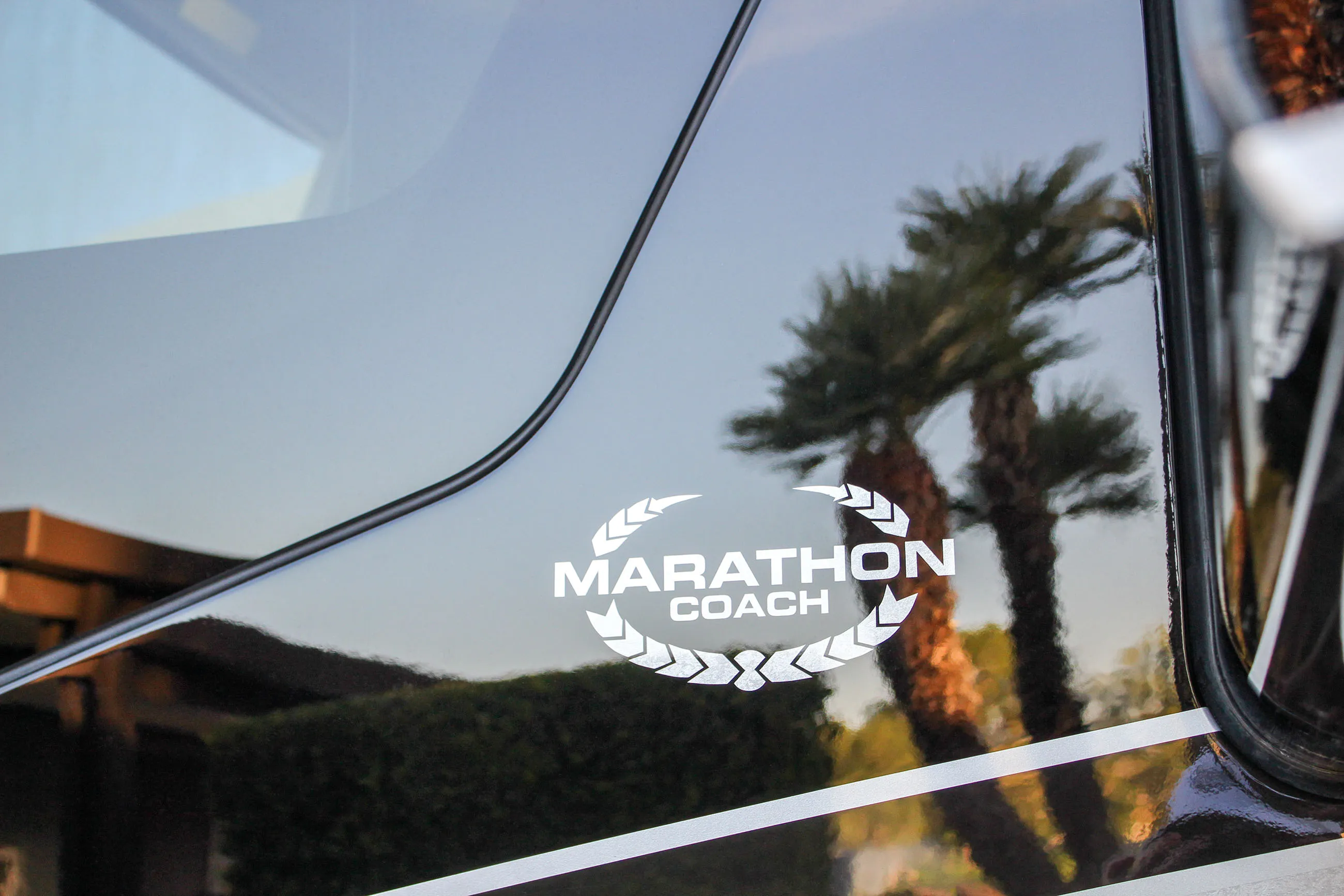 White Marathon Coach logo on a reflective black surface with palm trees and building reflected in it.