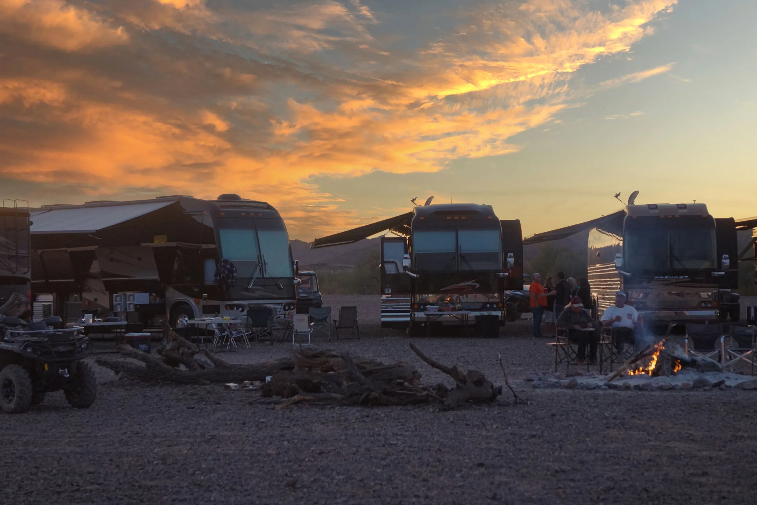 Several RVs with extended awnings parked on a gravel area at sunset, with people sitting around a campfire and socializing outdoors.