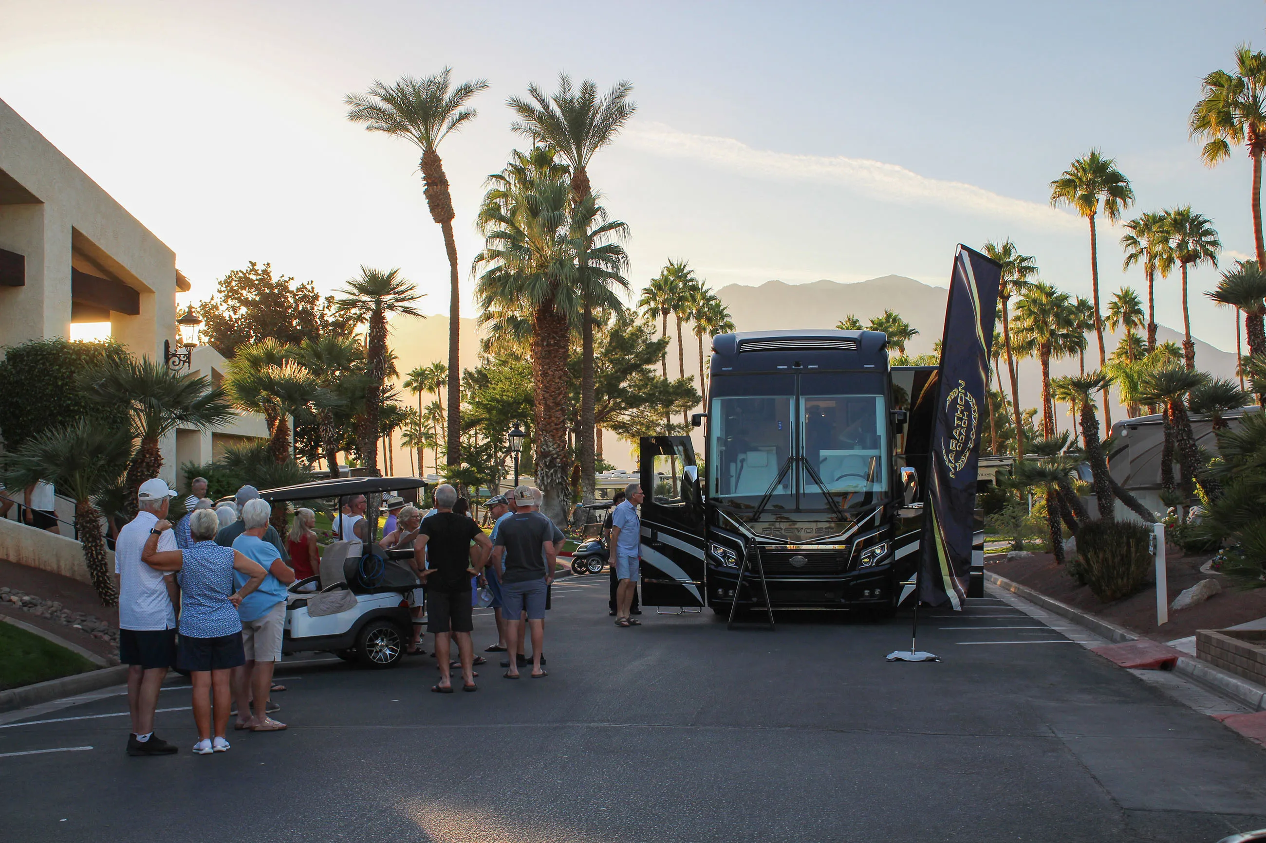 Group of people gathered near a black recreational vehicle and golf carts on a palm tree-lined street during sunset.