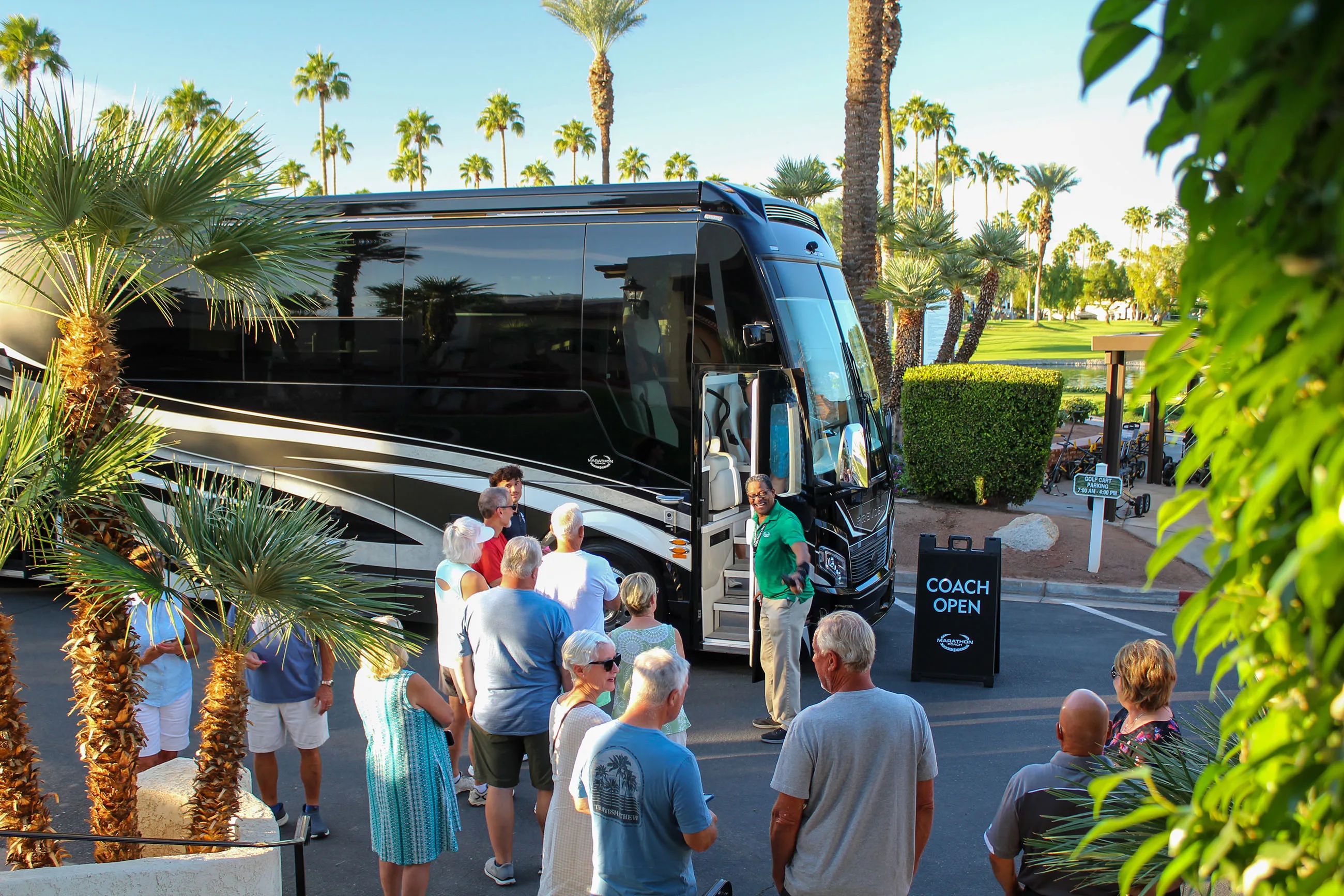 Group of people gathered near a black coach bus with a sign reading 'COACH OPEN' in a sunny, palm tree-lined parking area.