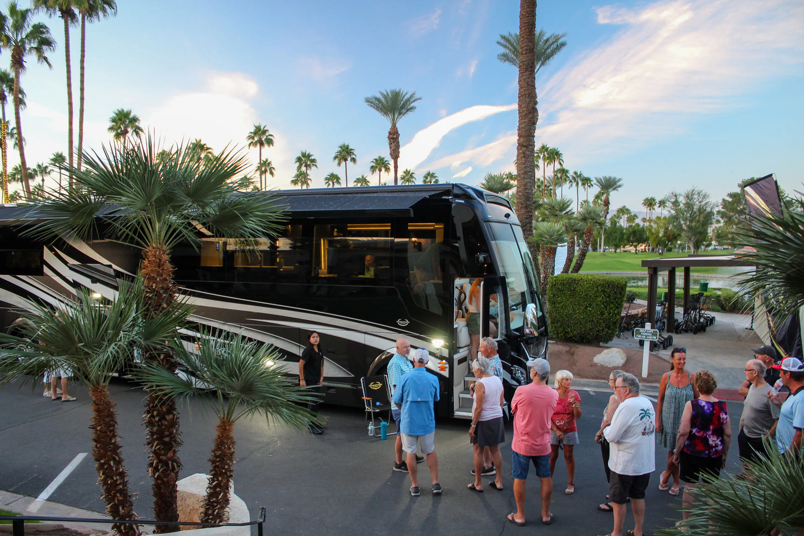 Group of people standing and chatting near a large black tour bus in a palm-lined parking area at sunset.