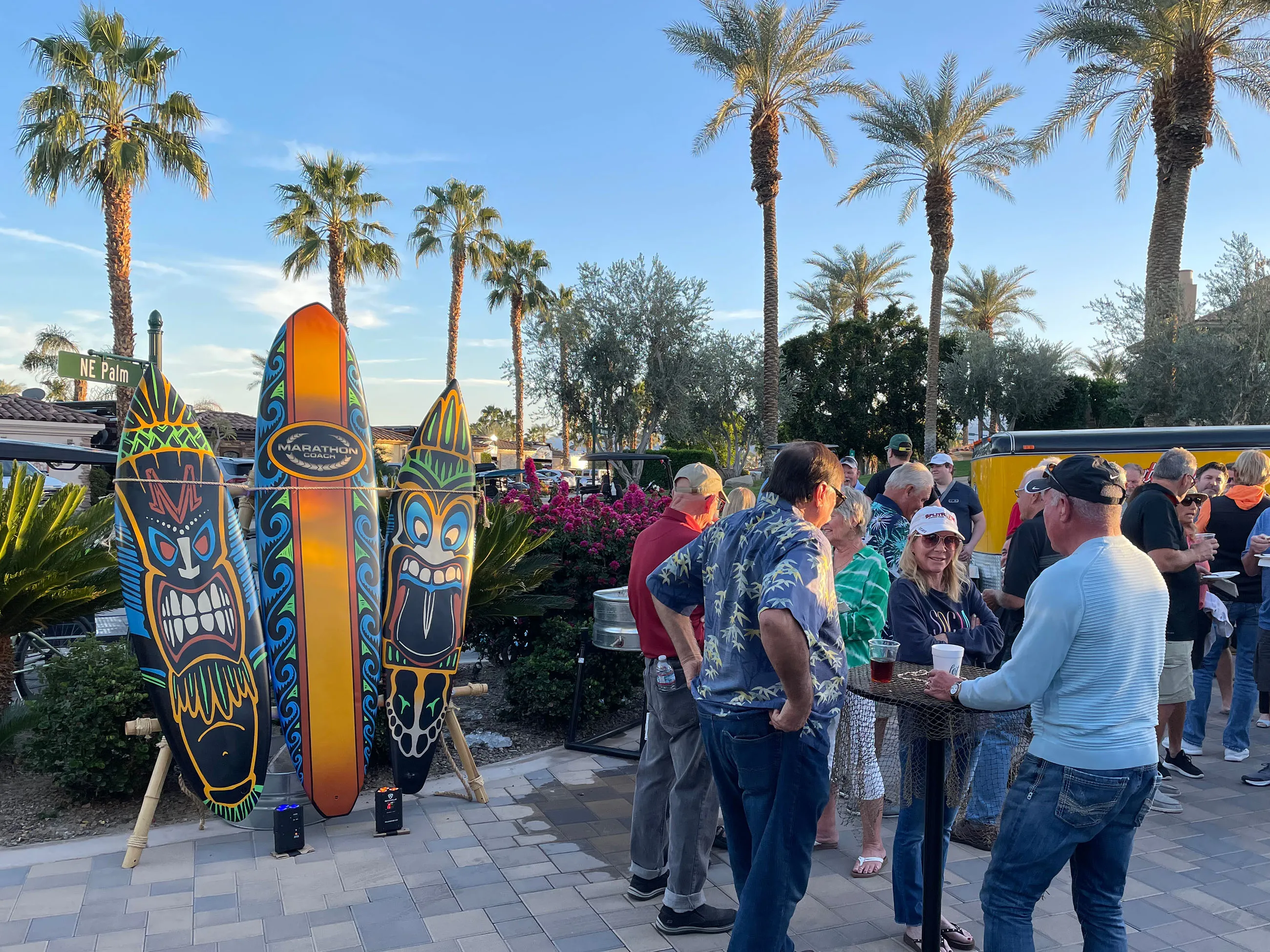 Group of people socializing outdoors near colorful tiki surfboard decorations under palm trees on a sunny day.