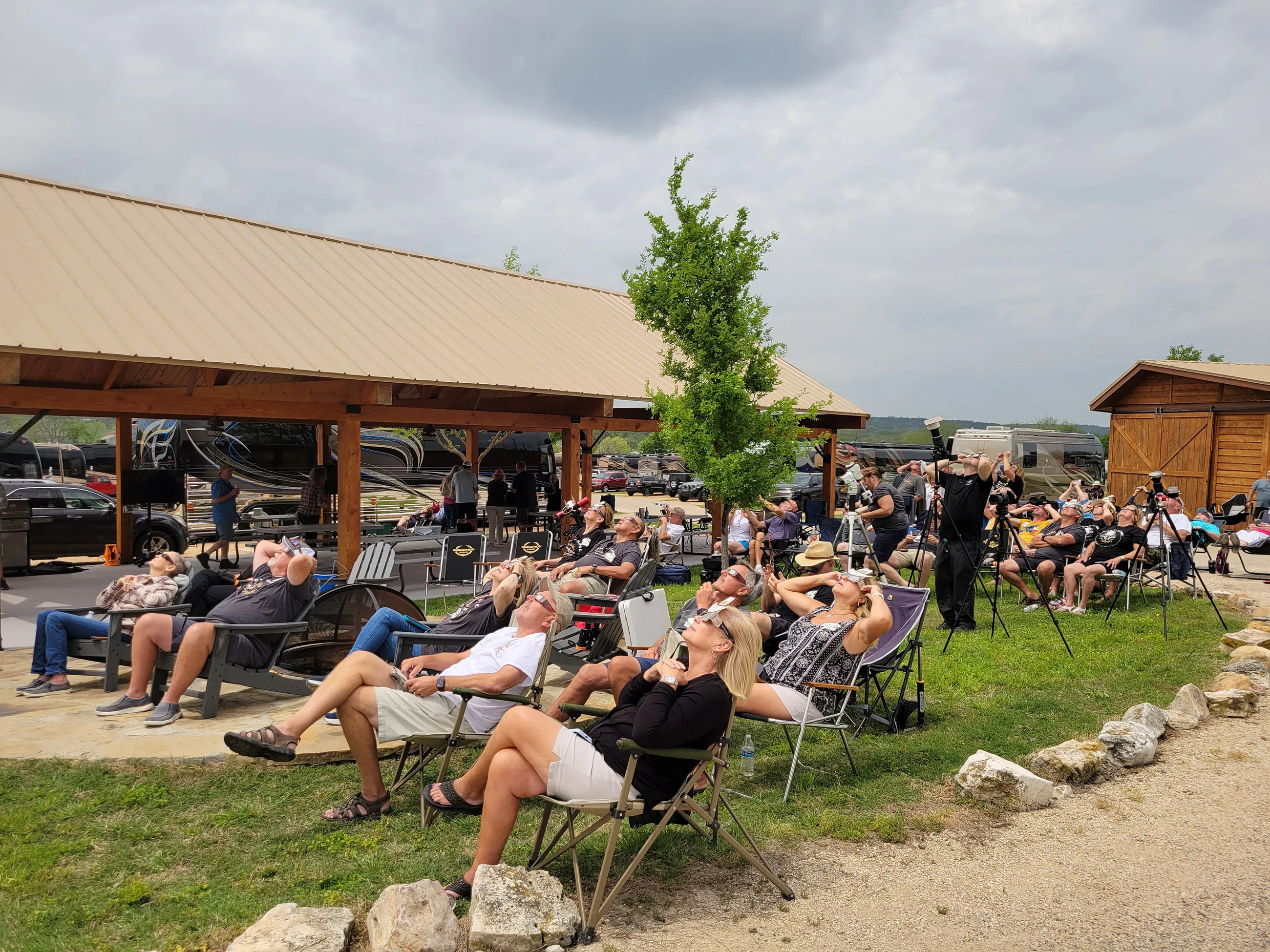Group of people sitting outside in chairs, wearing protective glasses and looking up at the sky, likely observing a solar eclipse.