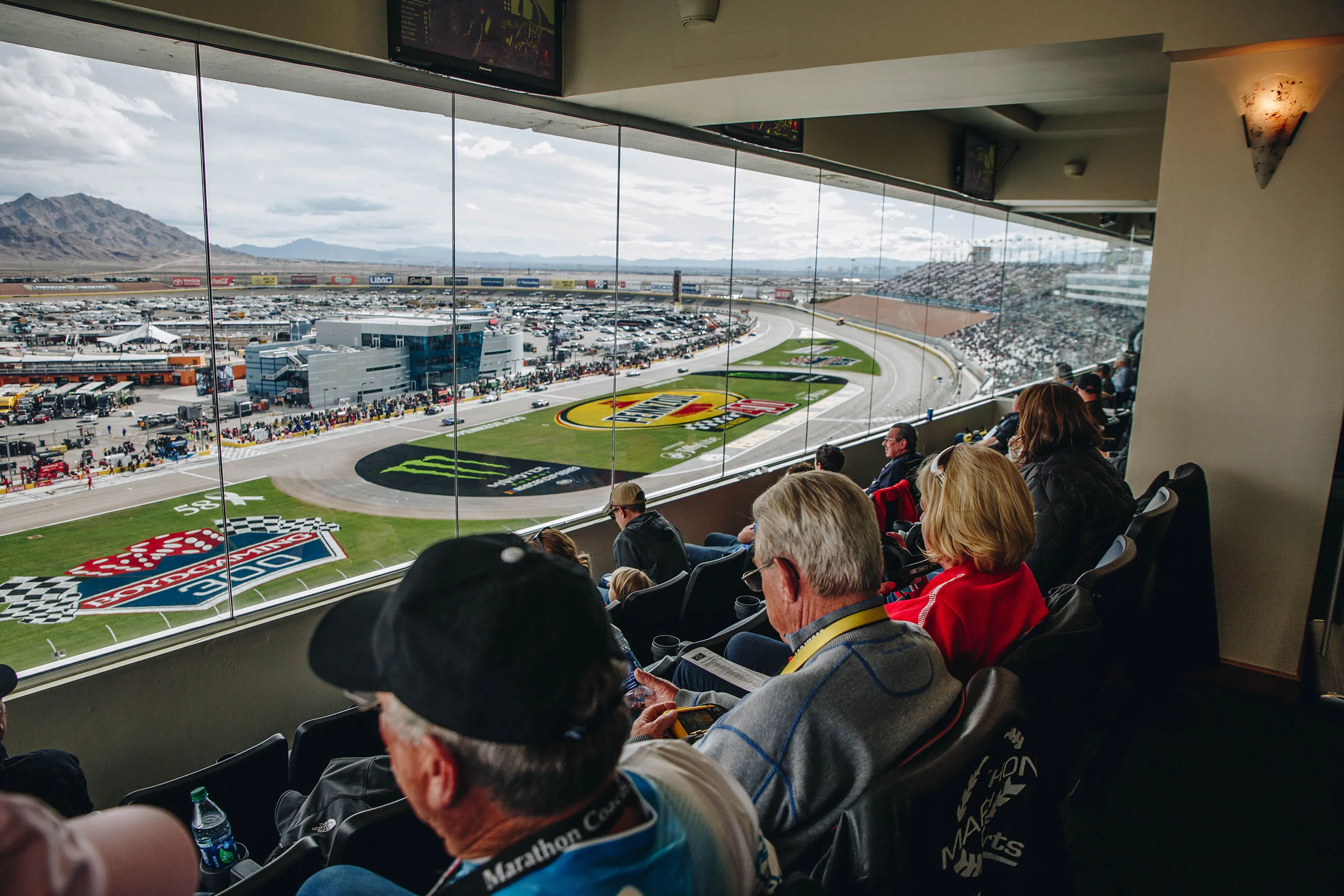Spectators watching a car race from a glass-enclosed suite overlooking a curved race track with sponsor logos on the grass.