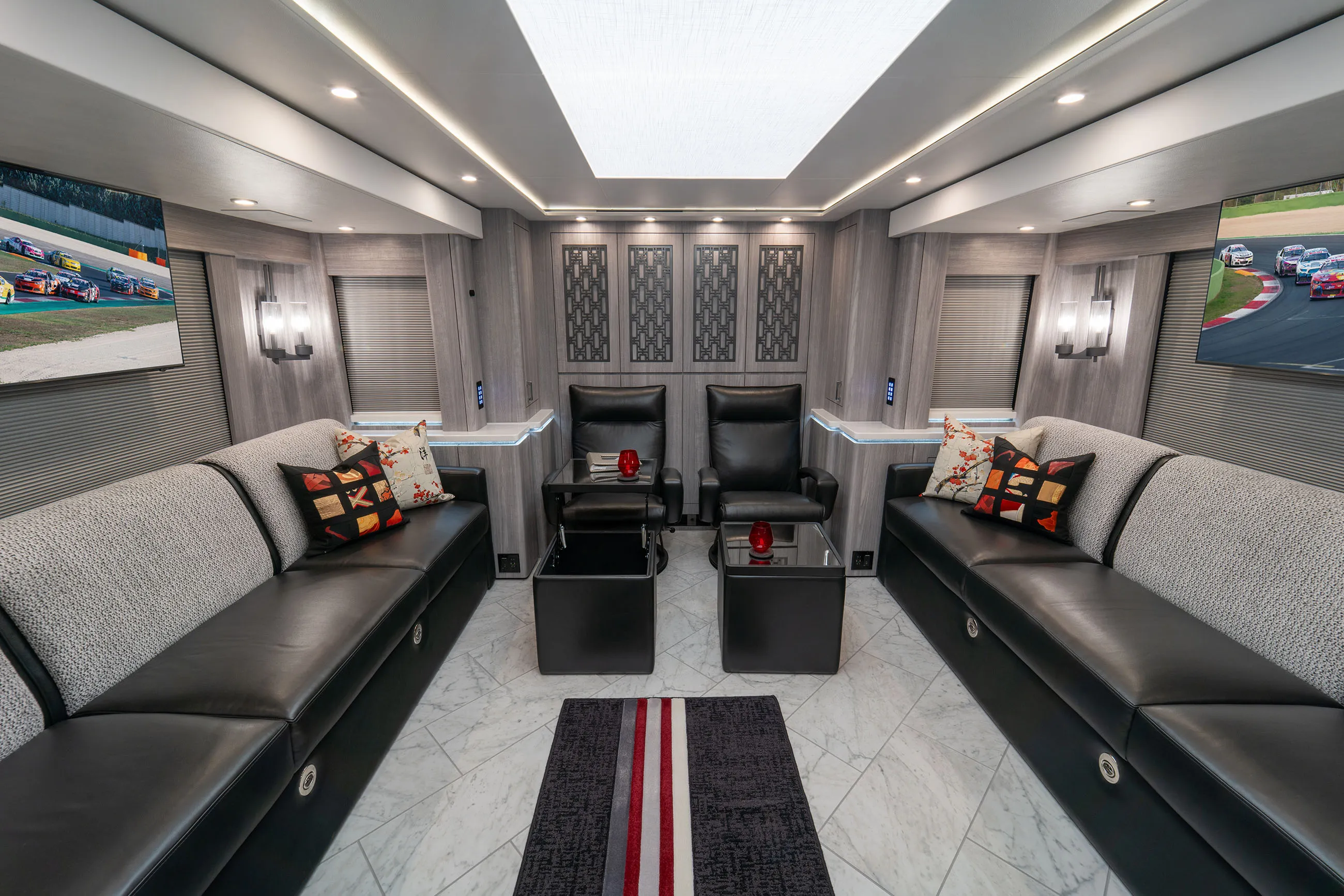 Modern RV interior featuring two long black leather sofas with patterned cushions, two black armchairs, two black side tables with red candles, decorative wall panels, and race cars shown on two mounted TVs.