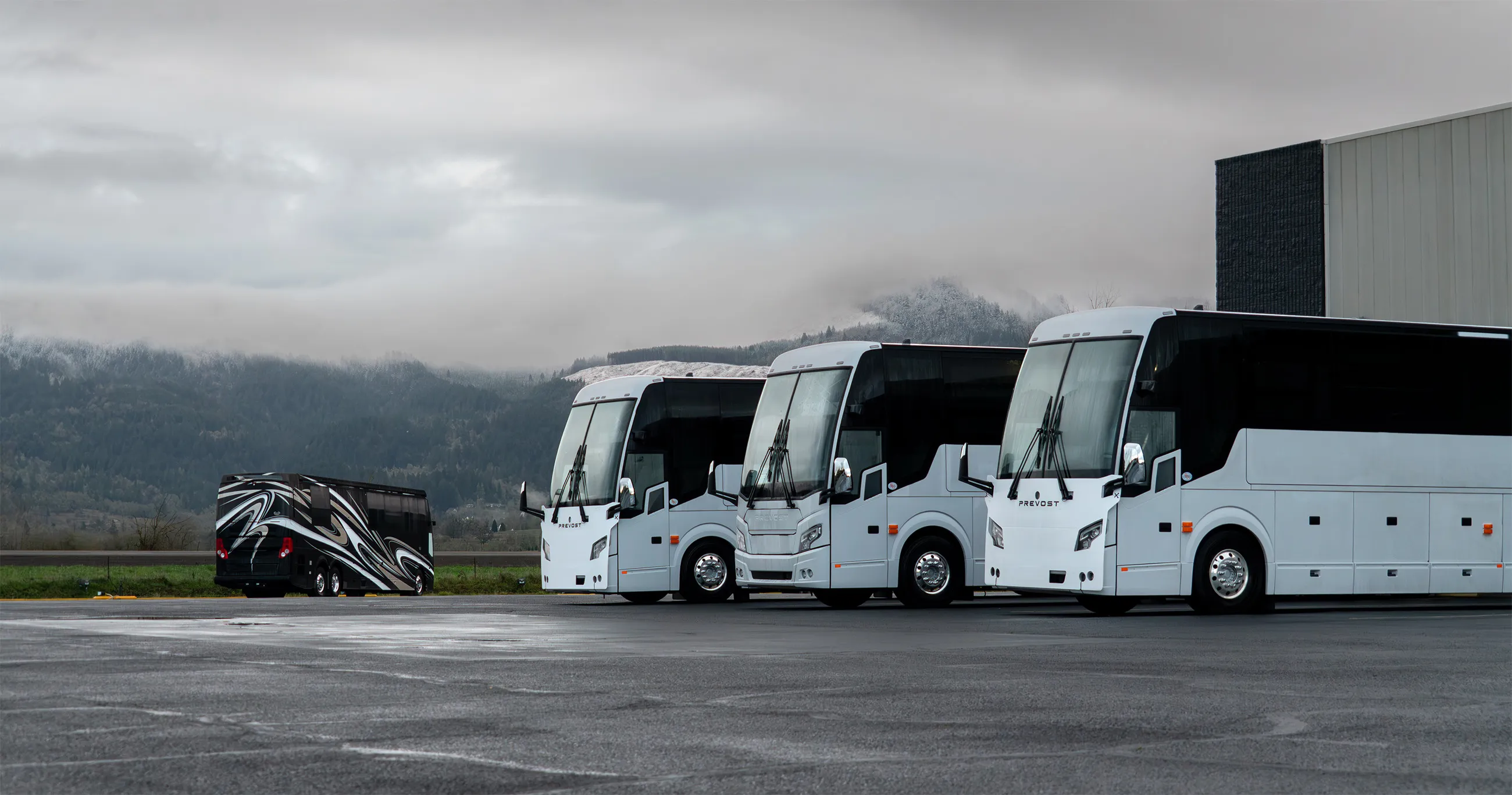 Three white Prevost chassis "shells" are ready for modification, while one black coach is parked farther back on a wet lot with foggy mountains in the background.