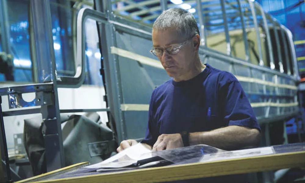 Engineer in safety glasses reading blueprints inside a bus frame assembly area.