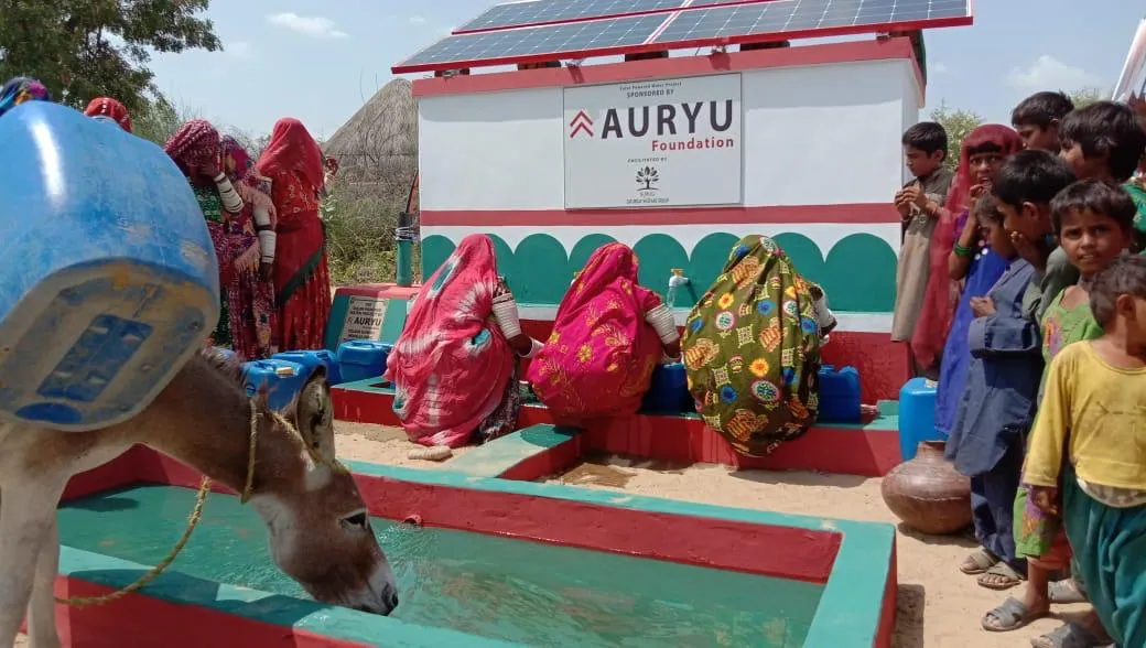 Donkey drinking water from a trough near women in colorful traditional attire filling containers at a solar-powered water station.