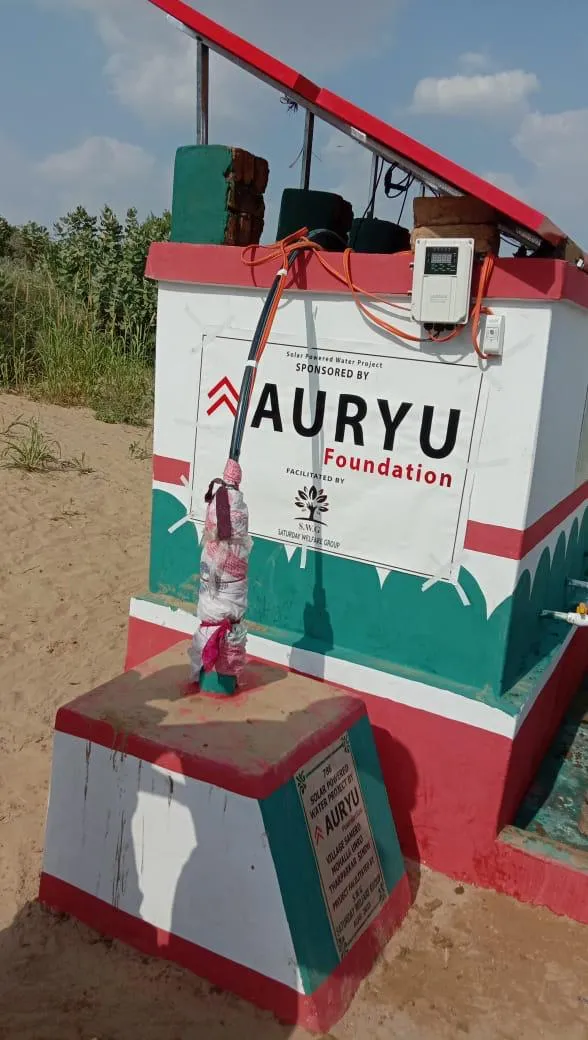 Solar-powered water pump installation with a sign showing sponsorship by Auryu Foundation in a sandy outdoor area.