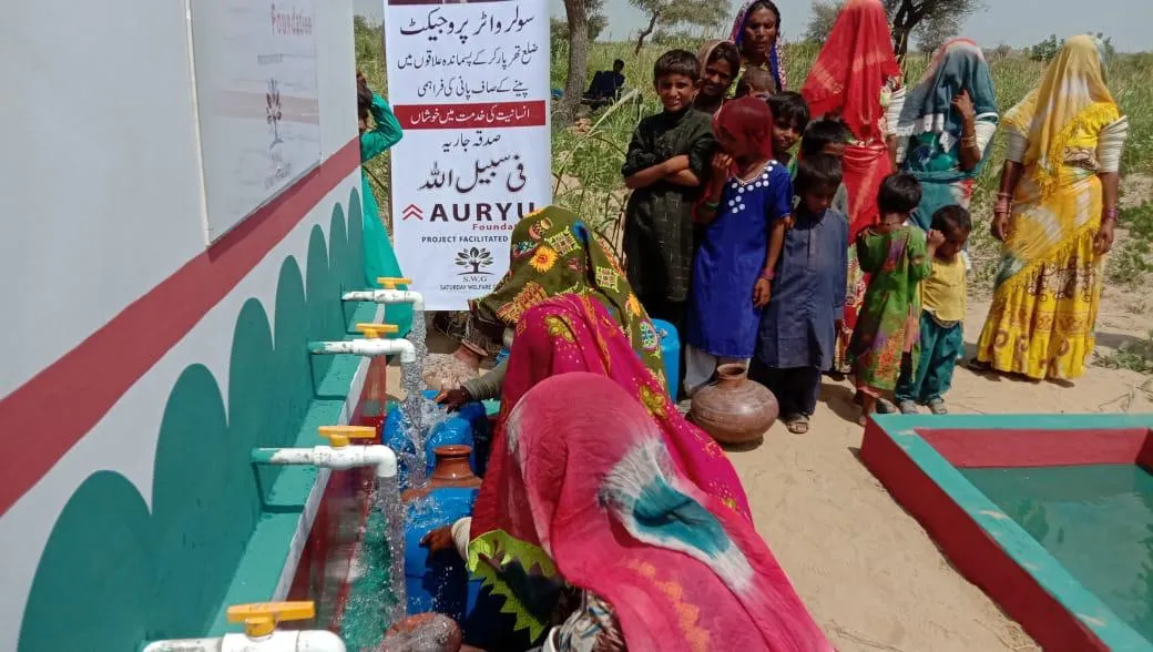 Women and children gather around multiple outdoor water taps, filling containers with water in a rural setting.