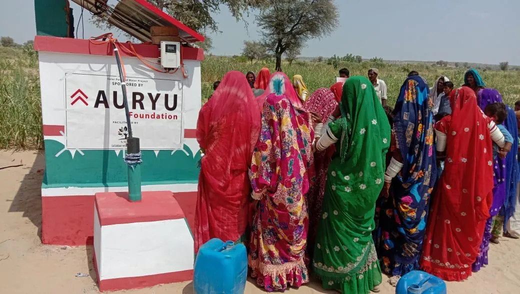 Group of women in colorful traditional clothing gathered around a water pump with a sign for Auryu Foundation in a rural outdoor setting.