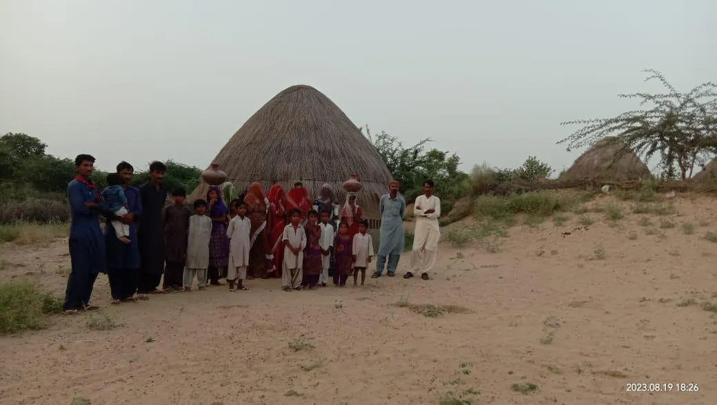 Group of men, women, and children standing in front of traditional thatched huts in a rural, sandy landscape.