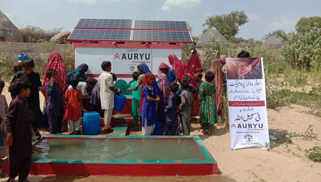 Group of children and women gathering water at a solar-powered water pump station with Auryu Foundation signage in a rural area.