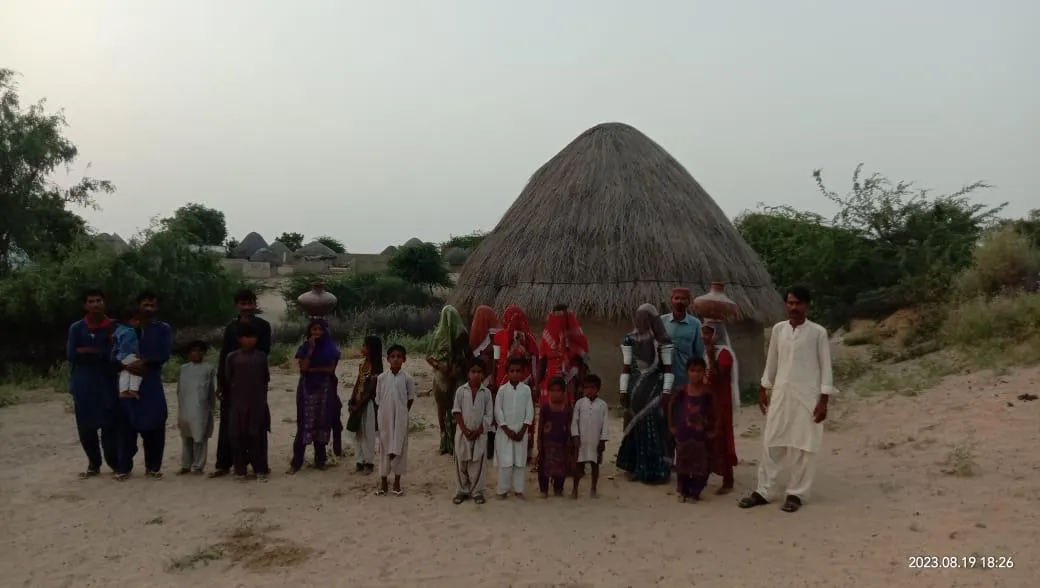 Group of men, women, and children in traditional clothing standing in front of a thatched round hut in a rural setting.