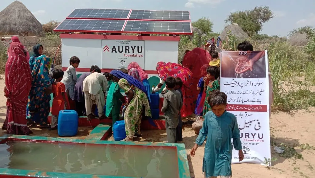 Group of people, including women and children, gathered around a solar-powered water pump station labeled AURYU Foundation in a rural area.