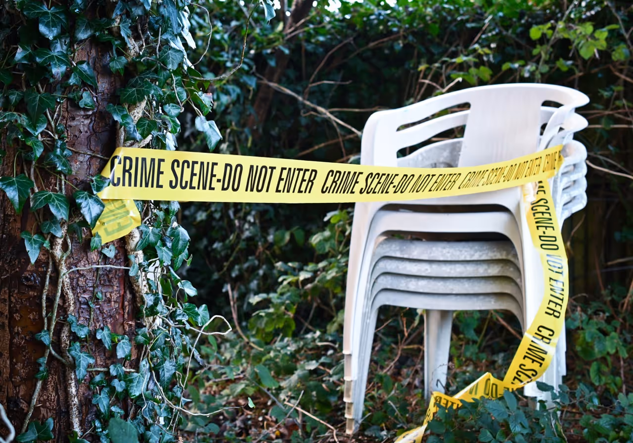 Stack of white plastic chairs wrapped with yellow crime scene tape in a wooded outdoor area.
