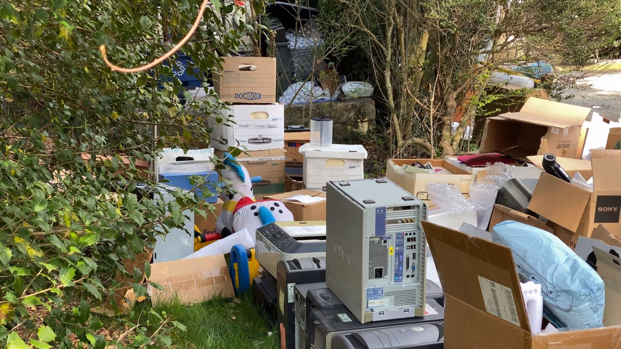 Outdoors cluttered with stacked cardboard boxes, old computers, a plush snowman toy, and various scattered items 