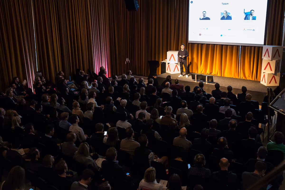 Large audience watch Antler representative provide presentation on stage with antler logo boxes next to him 