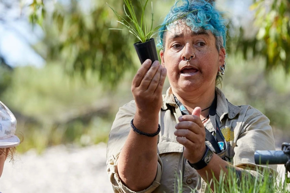 Garden educator holding a small native plant while explaining to a group outdoors.