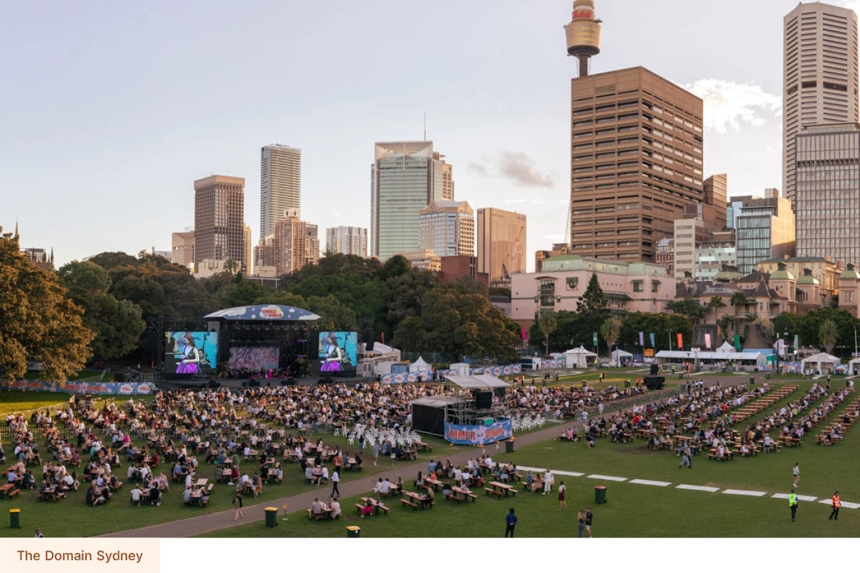 Large outdoor event at The Domain in Sydney, with crowds seated on the lawn and city buildings in the background.