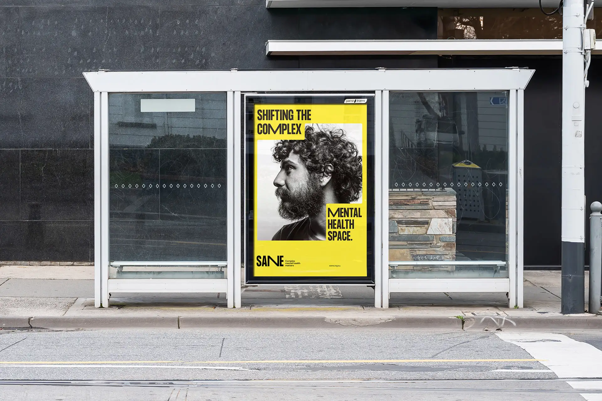A bright yellow SANE poster displayed at a bus stop, featuring a man in profile and the headline “Shifting the complex mental health space.”